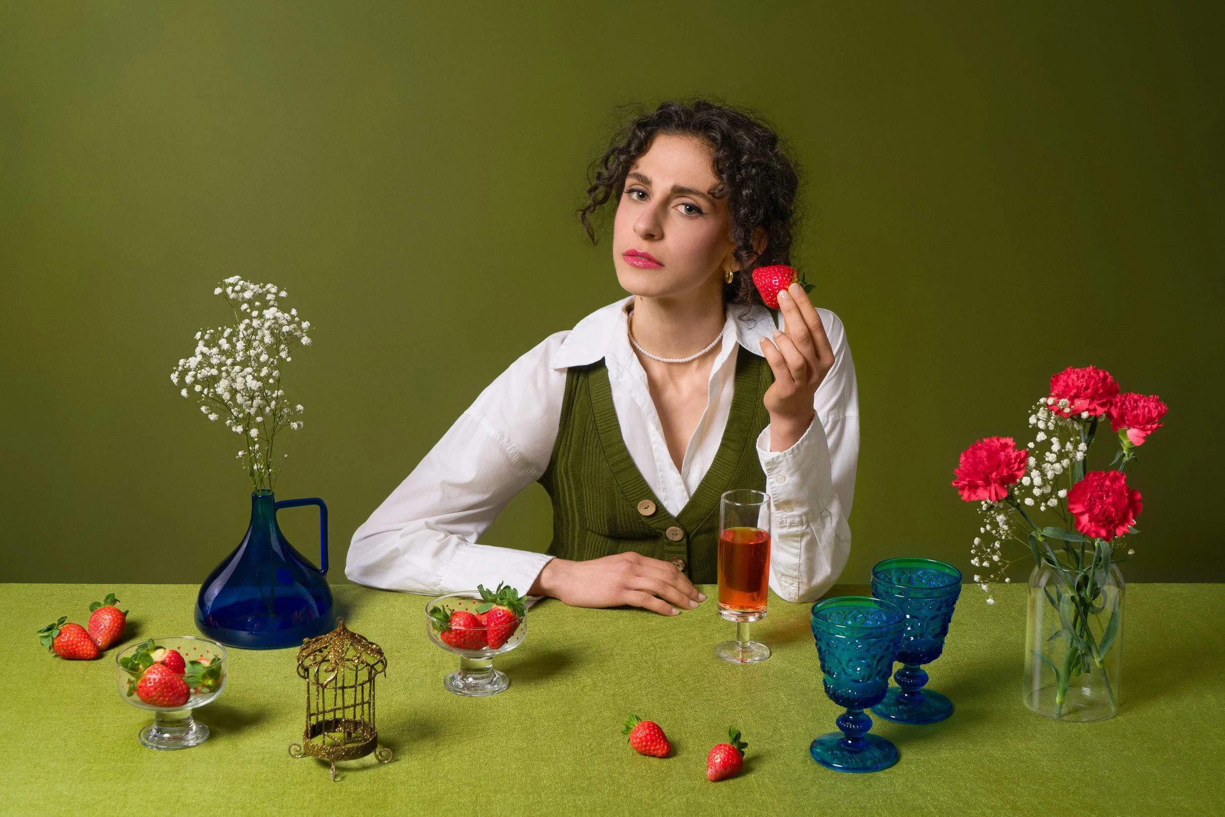 Una donna con capelli ricci e vestito a strati sorride e tiene una fragola vicino all'orecchio, circondata da fiori e stoviglie colorate su un tavolo verde.