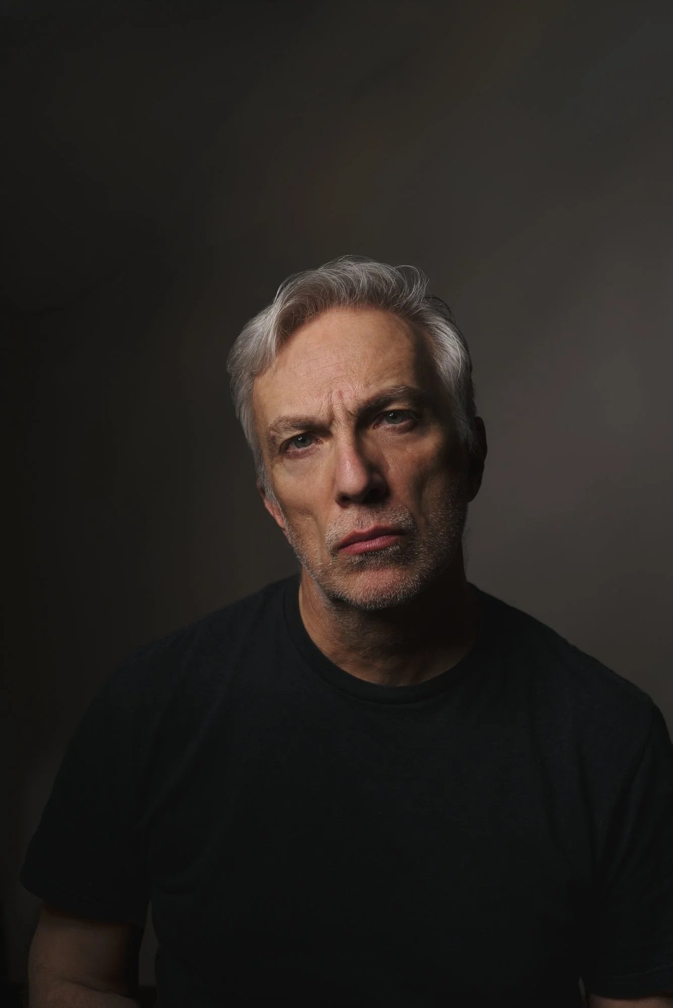 Ritratto di un uomo con capelli grigi, espressione seria, indossa una maglietta nera, sfondo scuro.