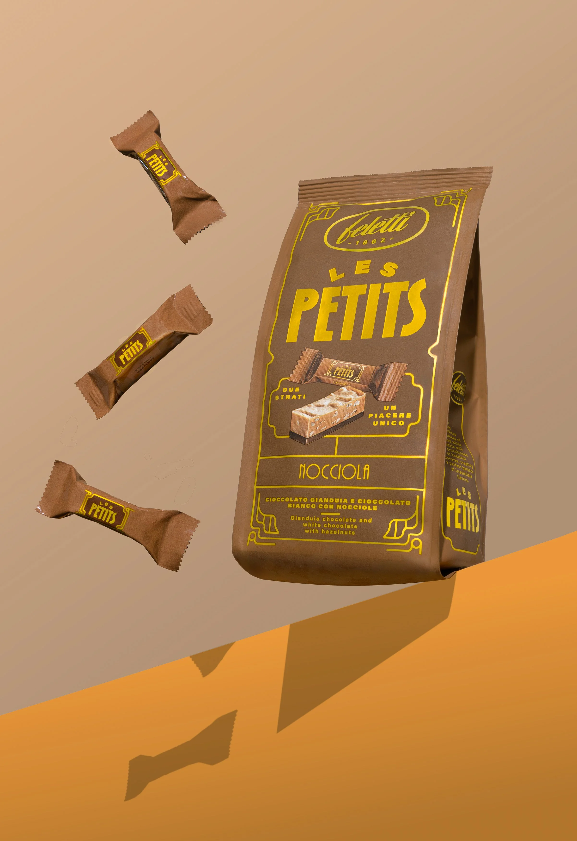 Confezione di cioccolato Les Petits di Feletti con nocciole e tre tavolette di cioccolato con nocciole che cadono, sfondo arancione e ombra proiettata alla confezione.