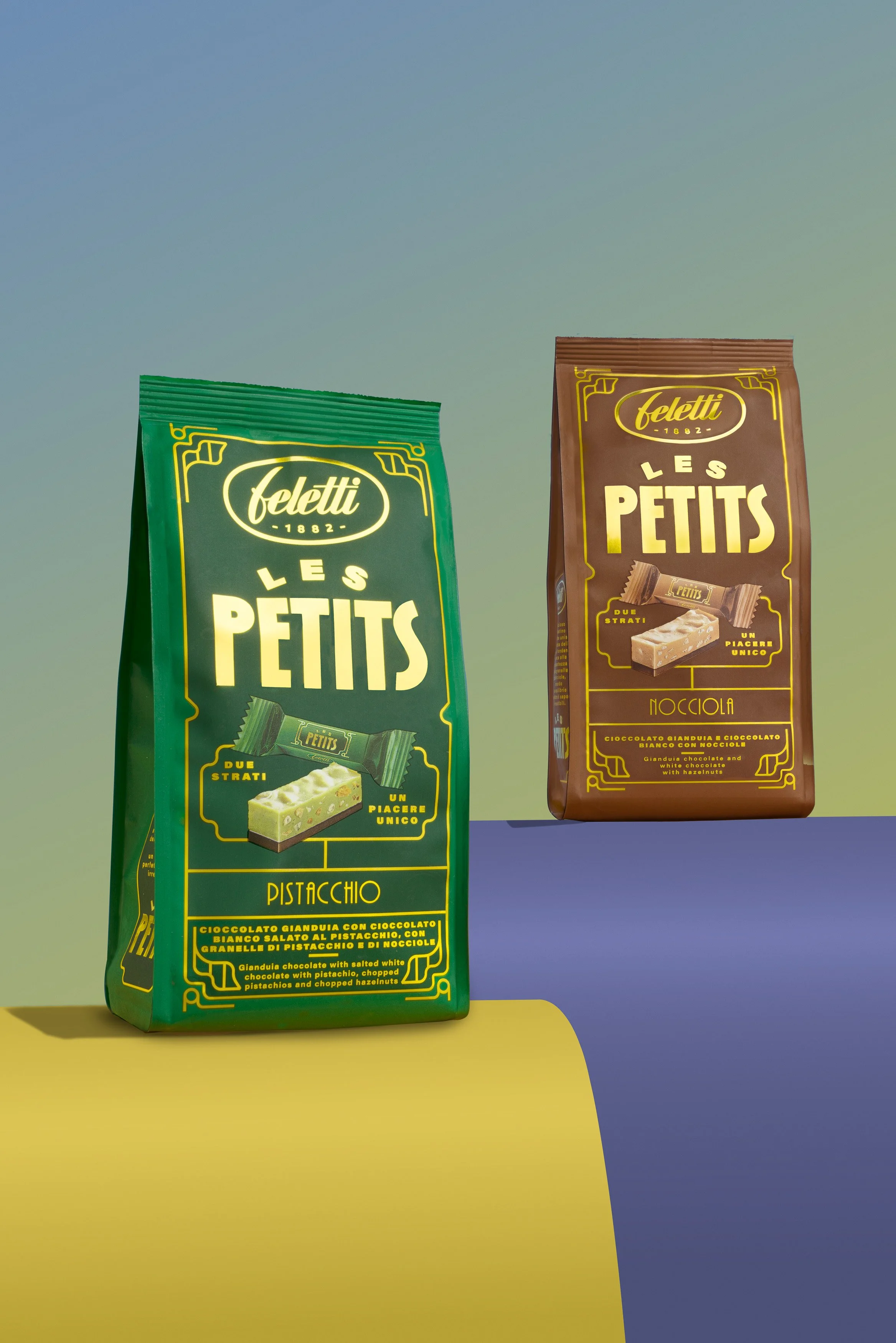 Due confezioni di cioccolatini Feletti Le Petits, una verde con pistacchio e una marrone con nocciola, su uno sfondo colorato con superfici gialle, blu e verdi.