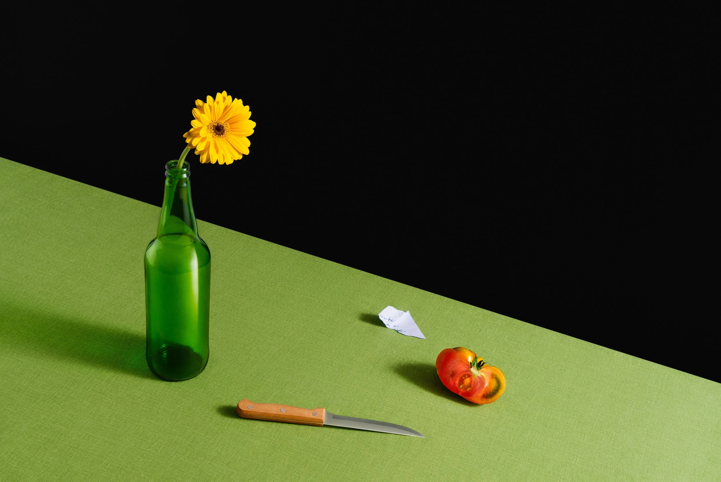 Una bottiglia verde con un fiore giallo in una sua apertura, su una tovaglia verde. Sul tavolo ci sono anche un foglietto di carta, un coltello da cucina e un pomodoro.