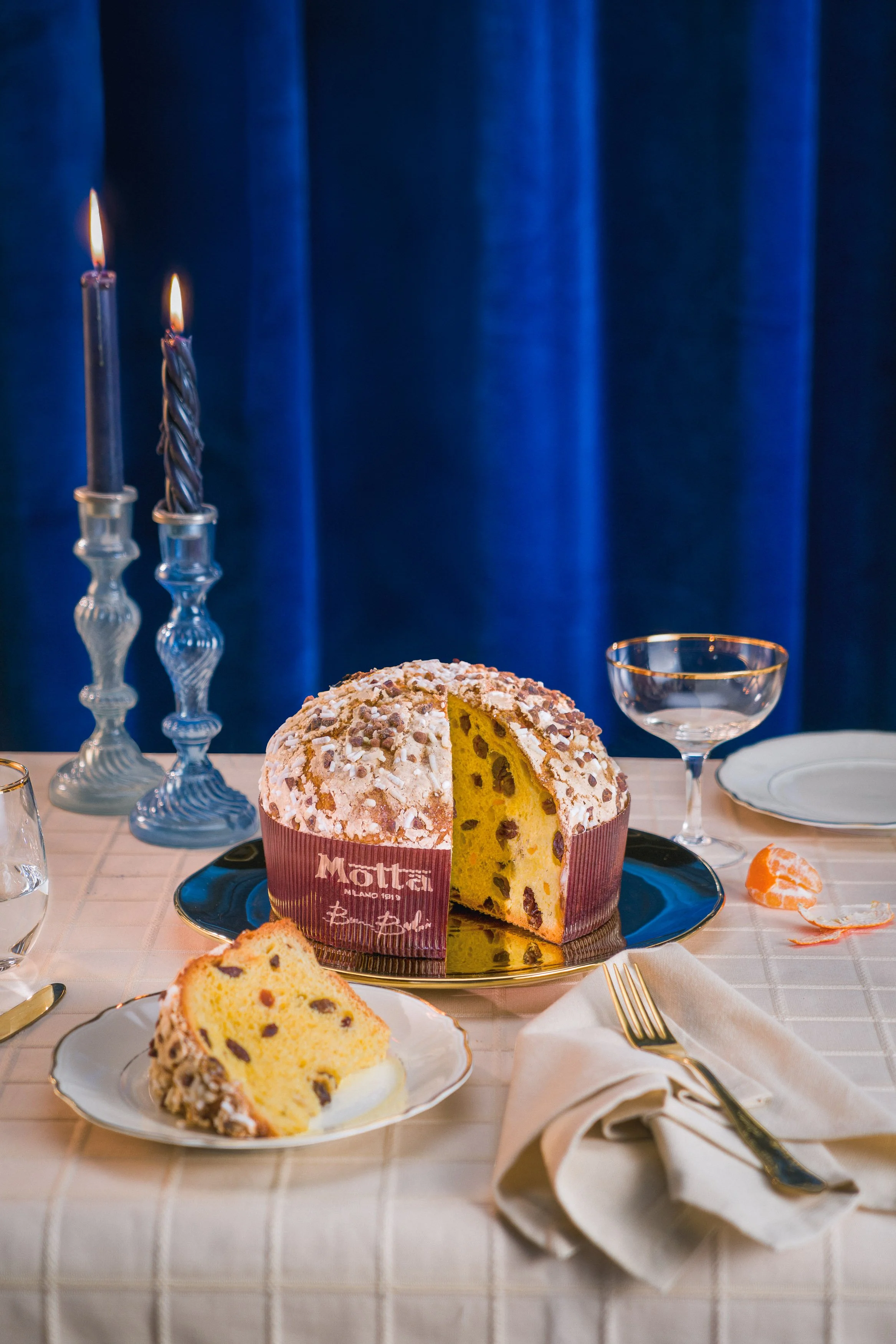 Torta di pan di Spagna decorata con zucchero a velo e canditi, situata su un piatto d'argento su una tavola imbandita con bicchieri e posate, con candelabri accesi e sfondo blu.