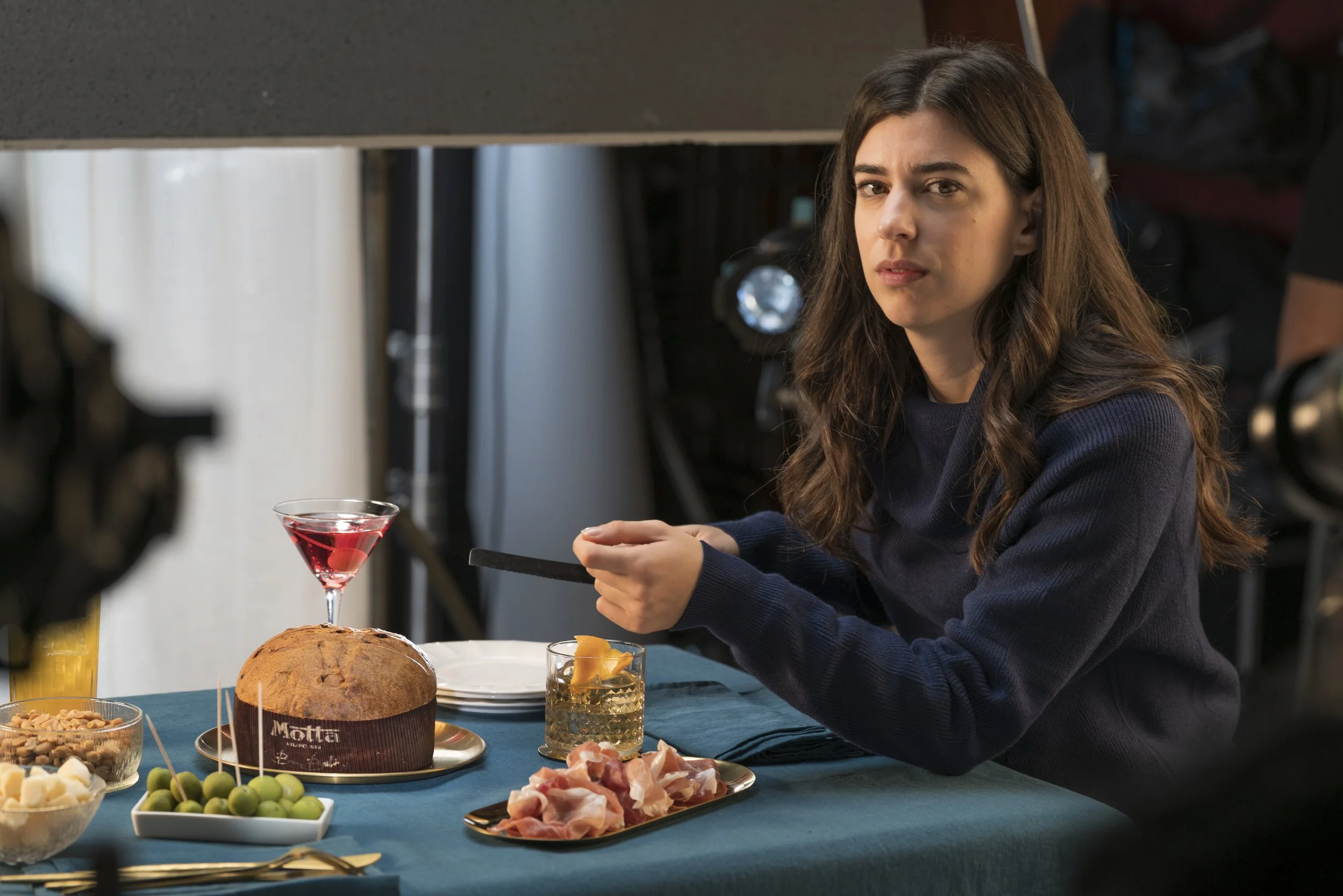 Donna seduta a tavola imbandita con cibo, tra cui pane, prosciutto, formaggi, olive, una torta e drink, in una scena di cucina o ristorante.