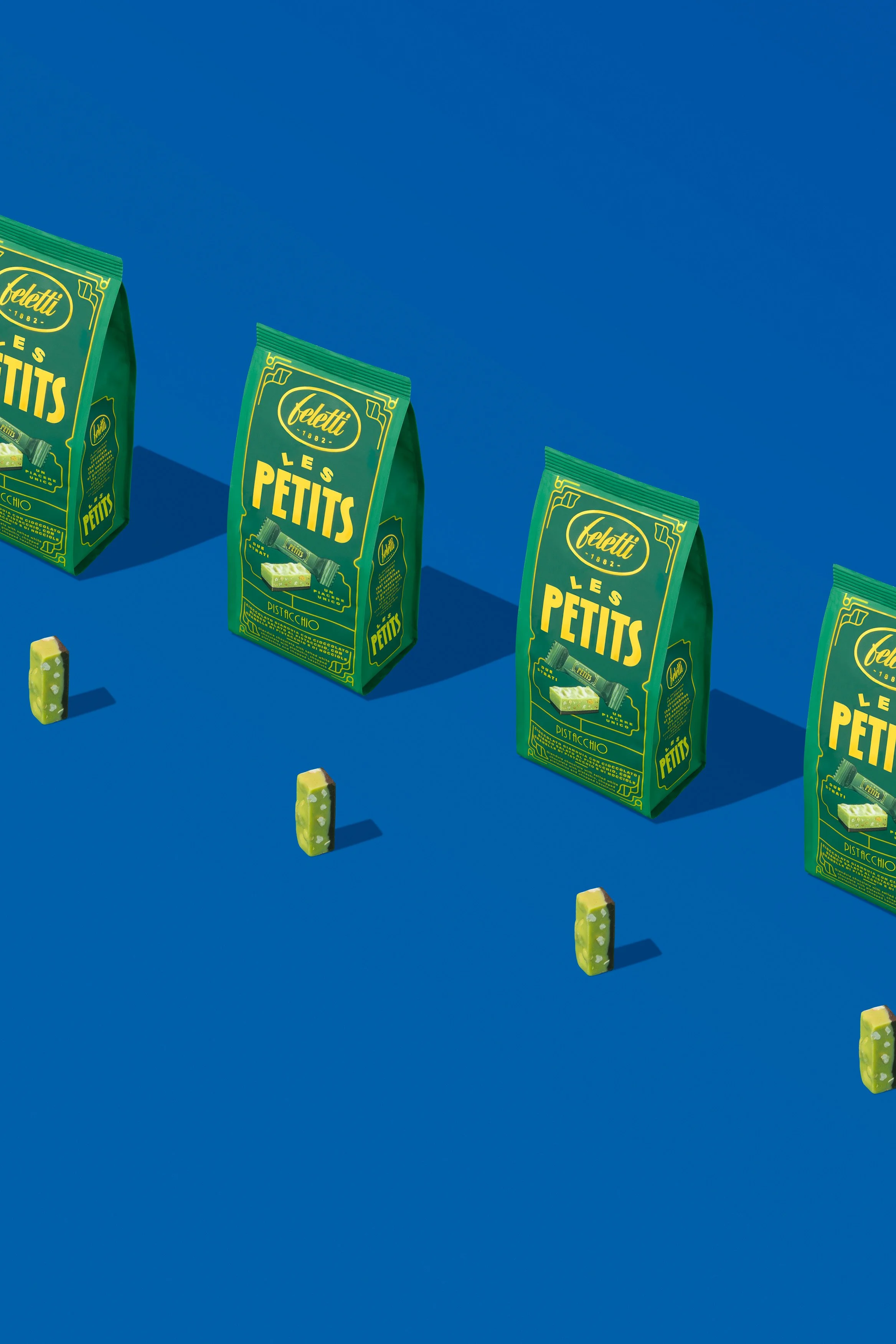 Pacchi di cioccolato verde con etichette gialle e dettagli decorativi, disposti in fila su sfondo blu con piccole caramelle piegate lungo la parte inferiore dell'immagine.