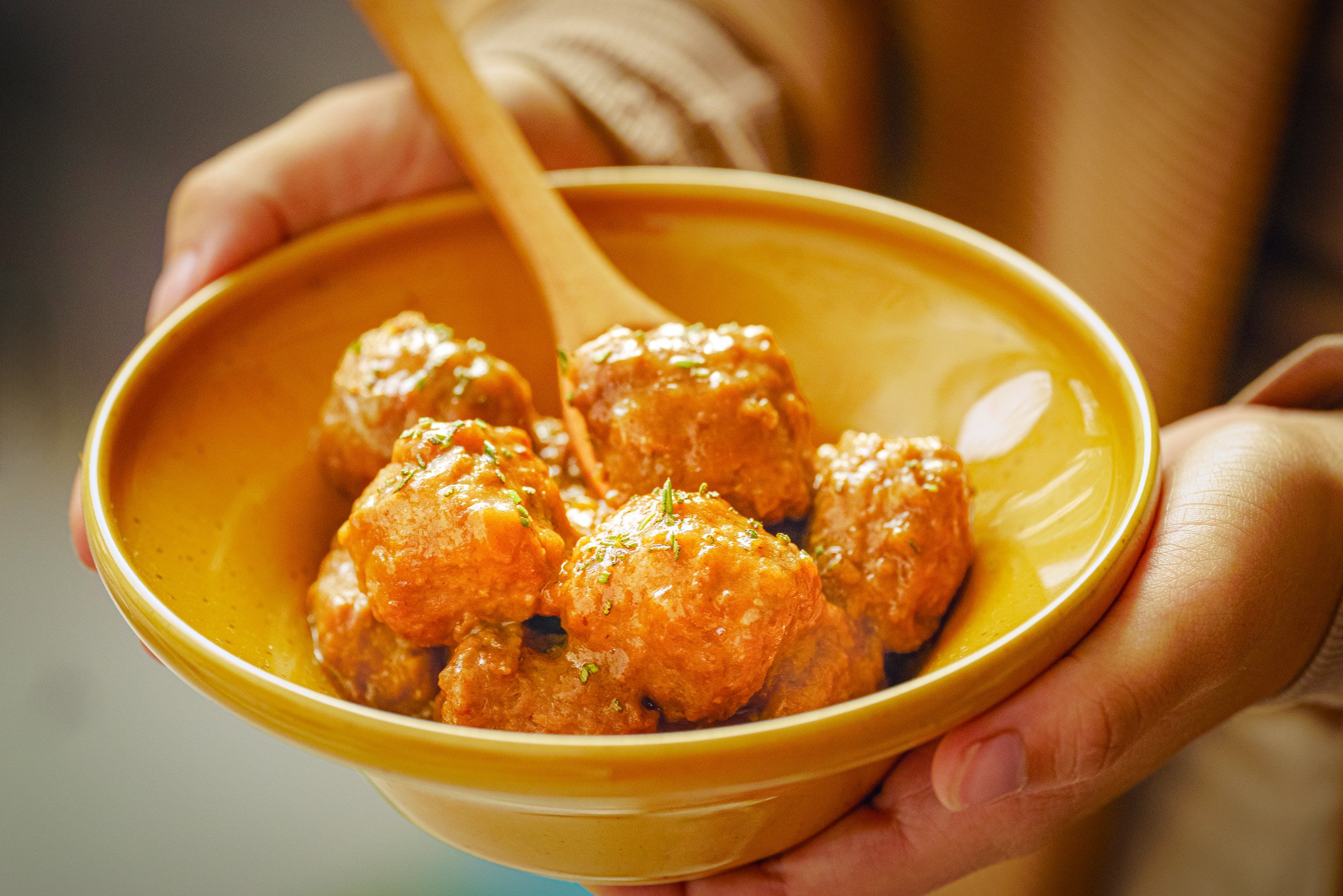 Piatti di polpette di carne in una ciotola gialla, con un cucchiaio, tenuto da una mano, in primo piano.