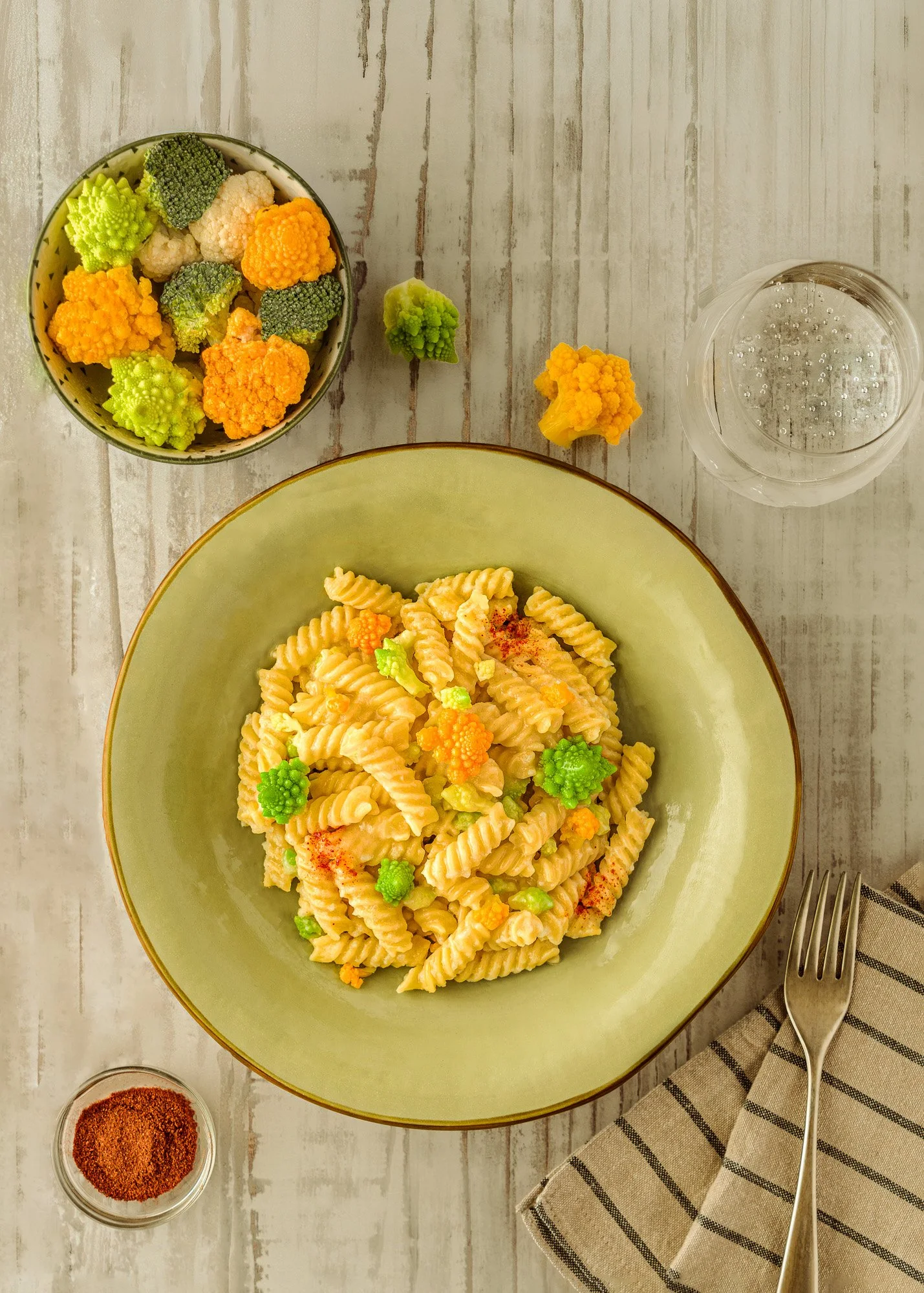 Piatto di pasta fusilli con cipolle e decorazioni di rapa raffa, con una ciotola di cavolfiore e broccoli colorati, un bicchiere d'acqua e un piattino con paprika in polvere, posato su tovagliolo a righe.