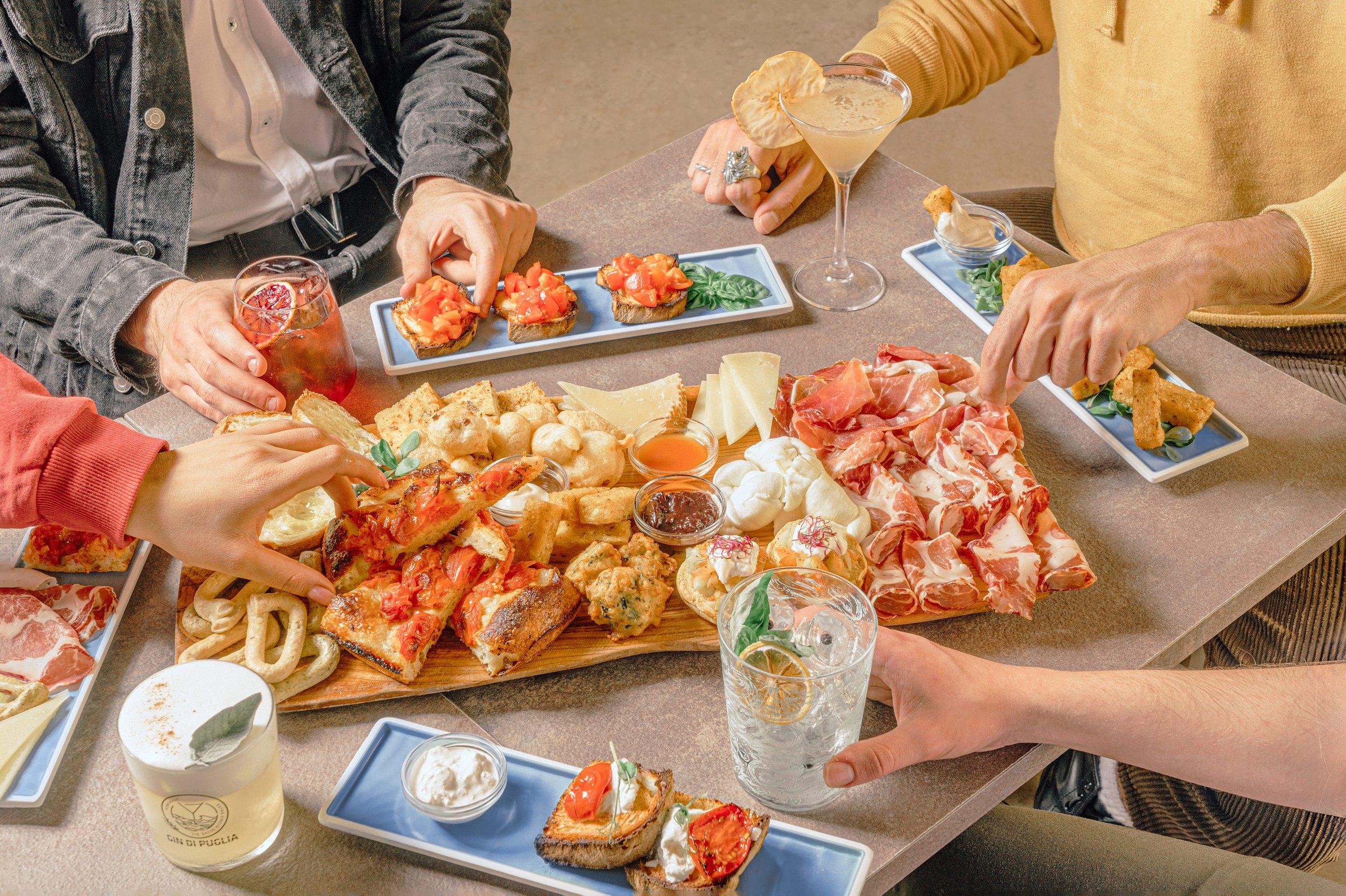 Persone che condividono un pasto con antipasti italiani, come salumi, formaggi, pizzerie e dolci, con bevande come cocktail e birra, su un tavolo in un ristorante