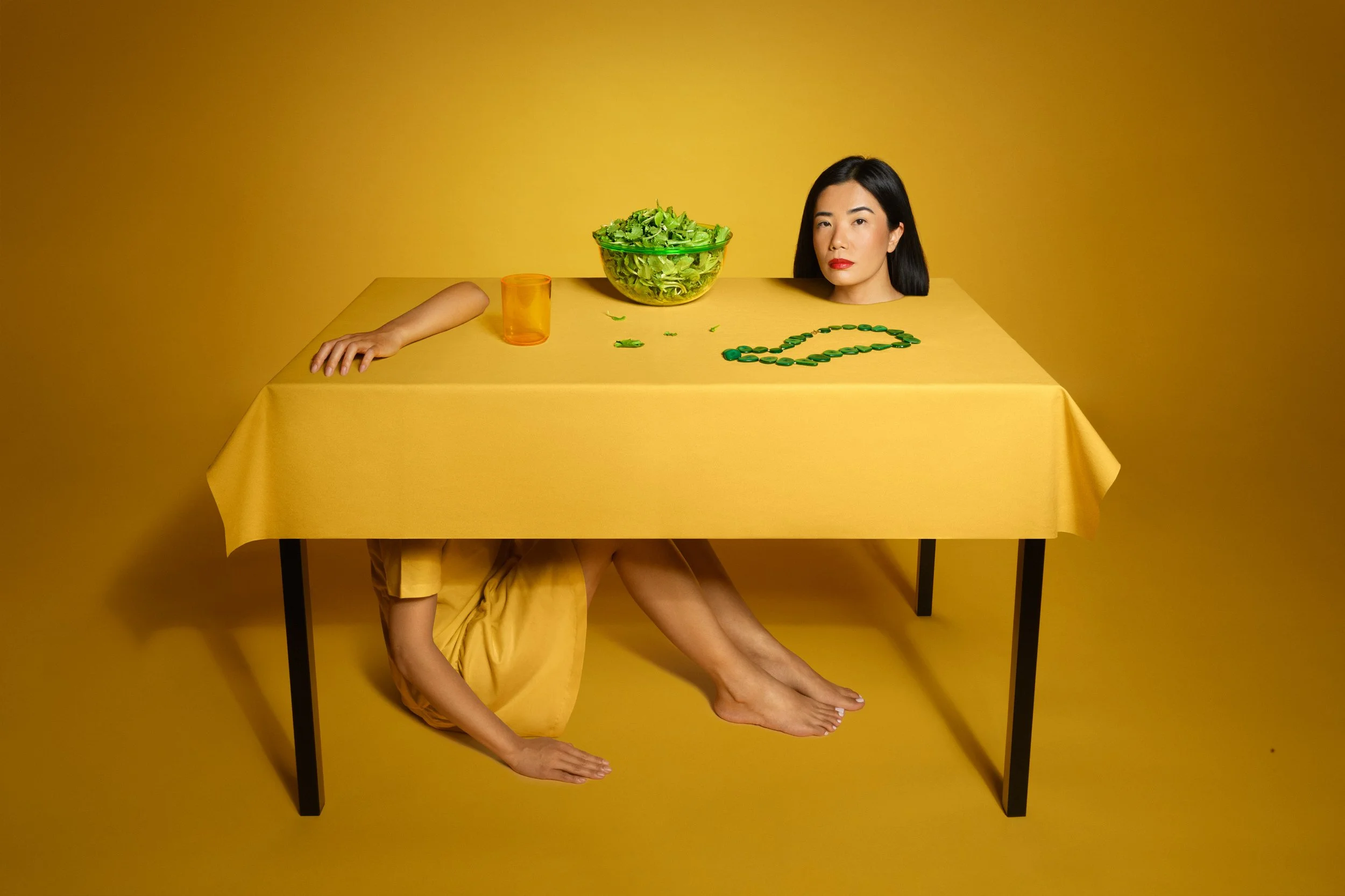 Una donna con i capelli neri e labbra rosse, con un braccio e una gamba appoggiati sul tavolo coperto di tessuto giallo, che si fonde con lo sfondo, e la testa che sporge di sopra, visibile solo la metà superiore del volto. Sul tavolo ci sono una gra