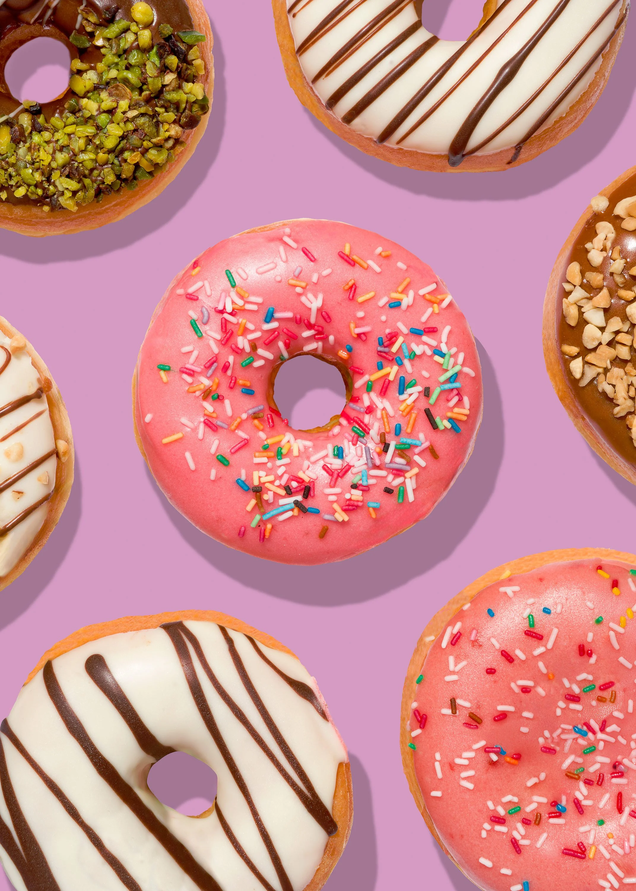 Diverse ciambelle decorate con glassa e zuccherini colorati, disposte su uno sfondo viola.