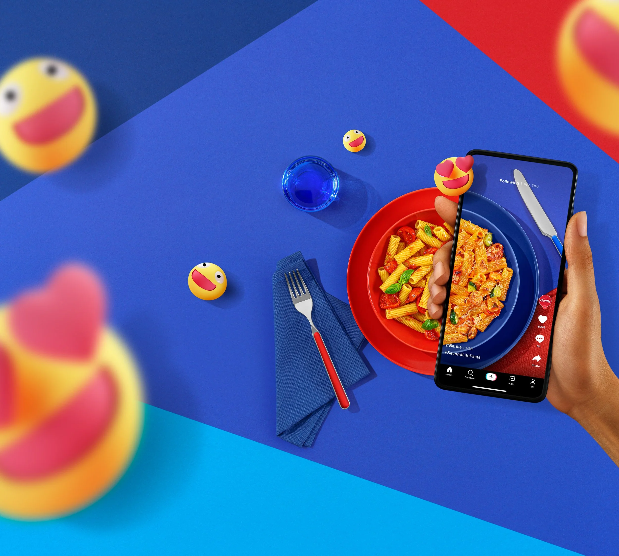Uno smartphone mostra un video con il logo di TikTok, posizionato sopra una ciotola rossa di pasta con pomodorini e basilico, su un tavolo blu con posate e vetro d'acqua, decorato con emoji di faccine sorridenti con occhi a cuore.