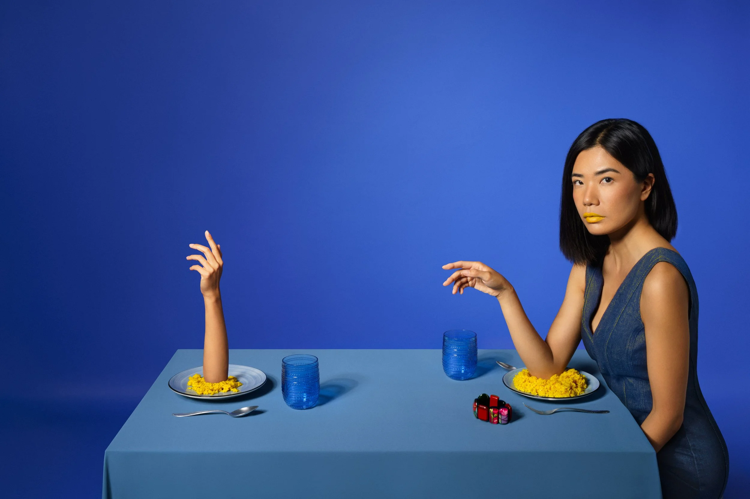 Donna seduta a un tavolo blu con due piatti di riso giallo e due bicchieri azzurri, con un uno che ha il braccio mozzato e la mano che si alza come per saluto.