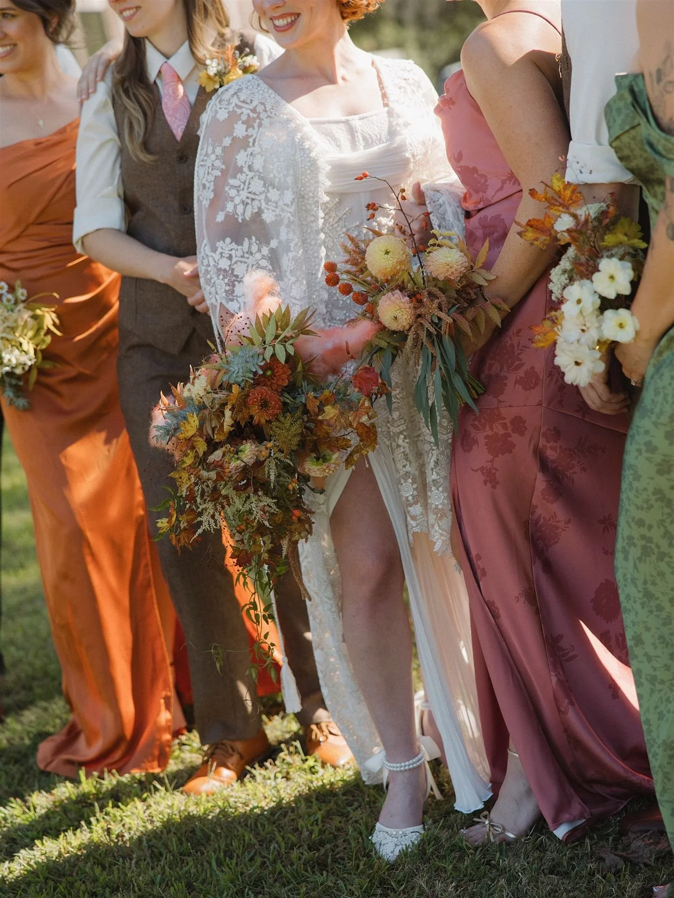 Mismatched bridesmaids dresses in fall colors, orange, brown, green, rosy pink. Bride in a bridal cape and high slit couture bridal dress by wedding planners epoch co