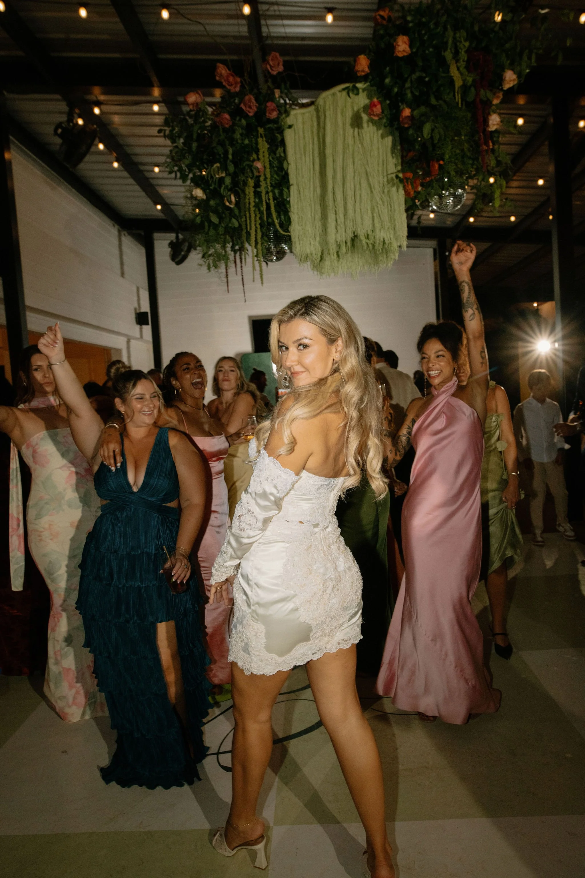 Bride in mom's wedding dress remade into short lace reception dress dancing with friends under floral install — playful, luxurious Gen Z wedding energy