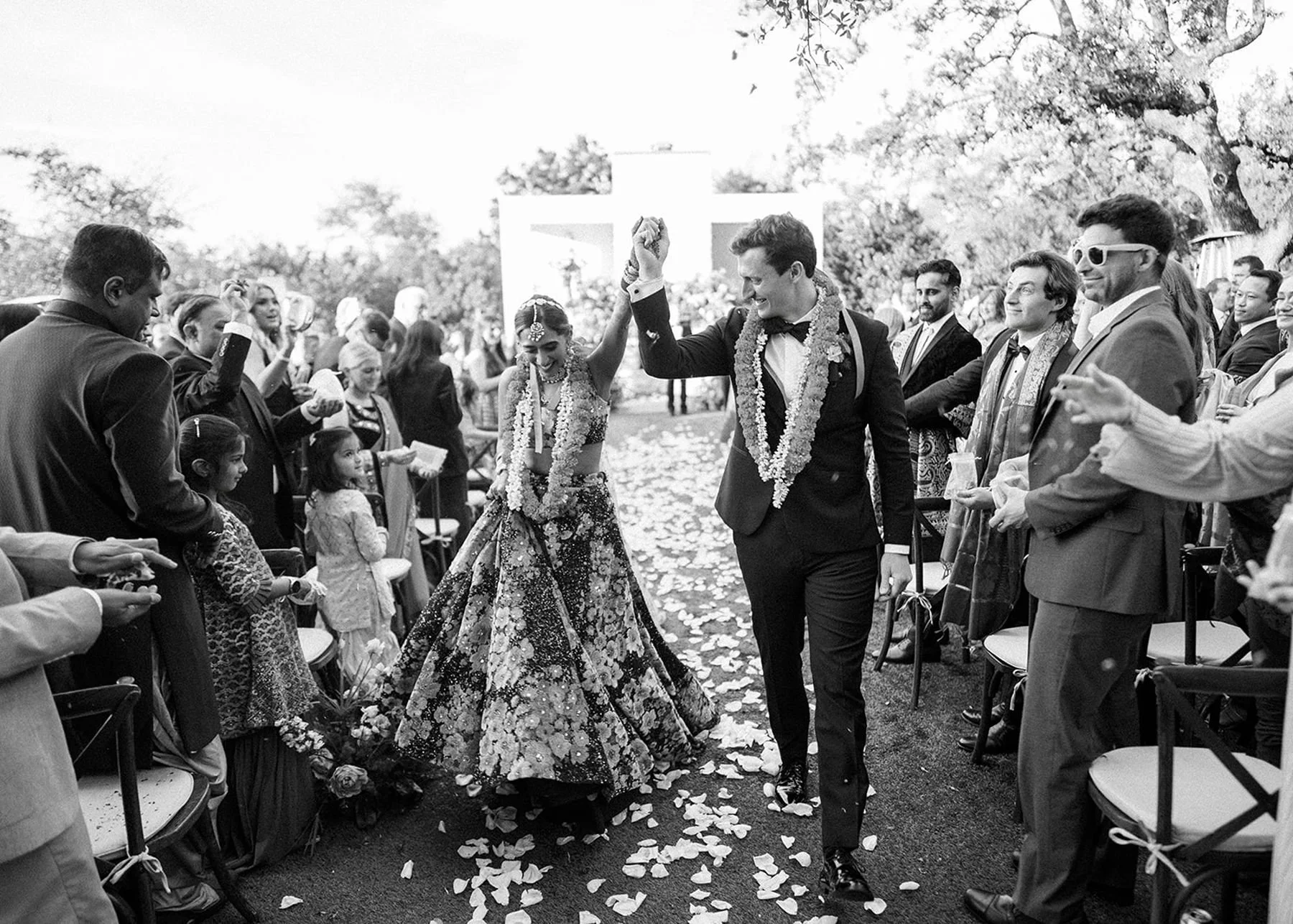 Bride and groom holding hands and celebrating their wedding as they walk down the aisle lined with petals and guests celebrating with them - black and white ceremony picture at the arlo wedding venue wedding planners wedding planners
