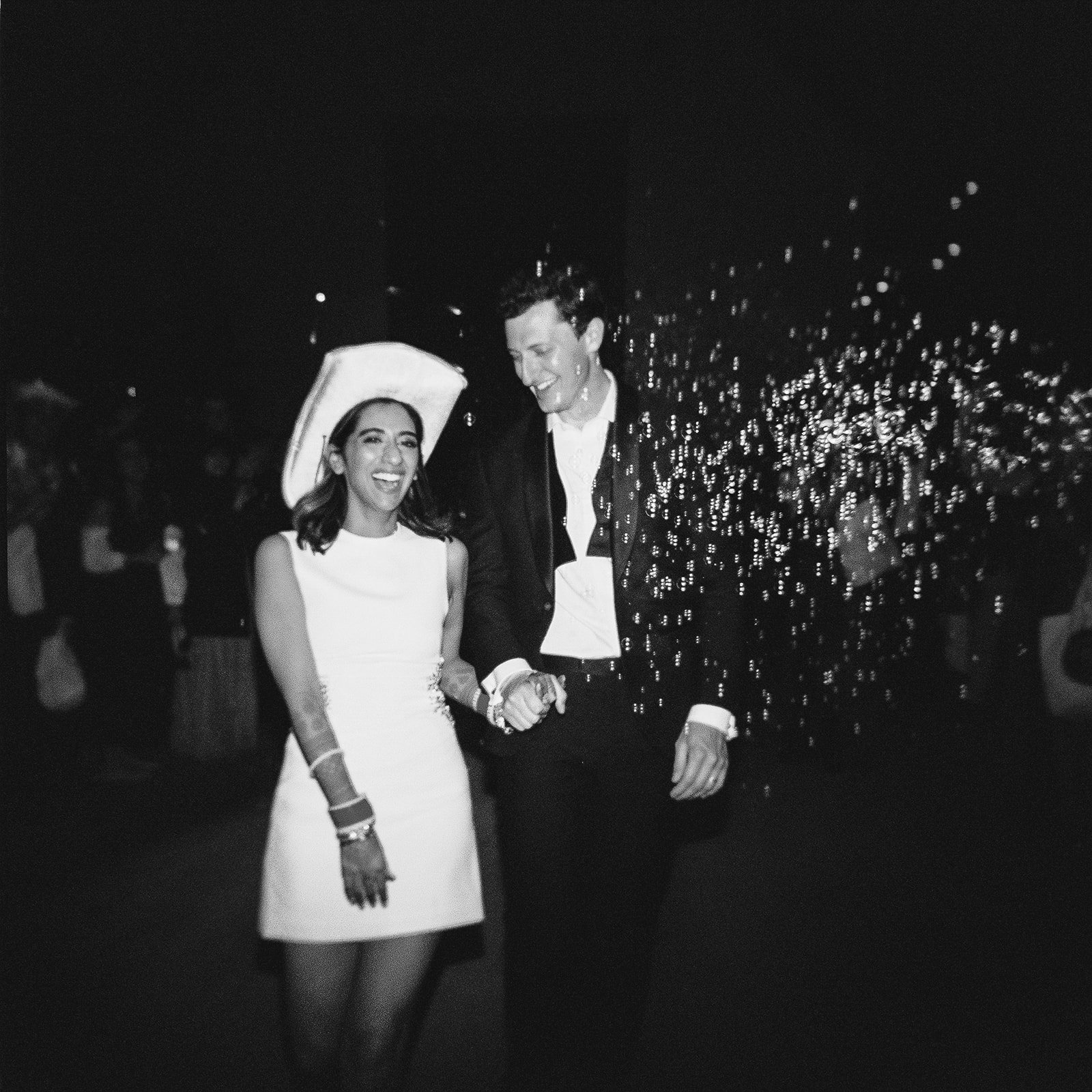 A groo and bride holding hands and smiling at each other at a nighttime wedding exit. The bride is wearing a sleeveless white dress and a led cowgirl hat; the man is dressed in a dark suit with an open-collared shirt.