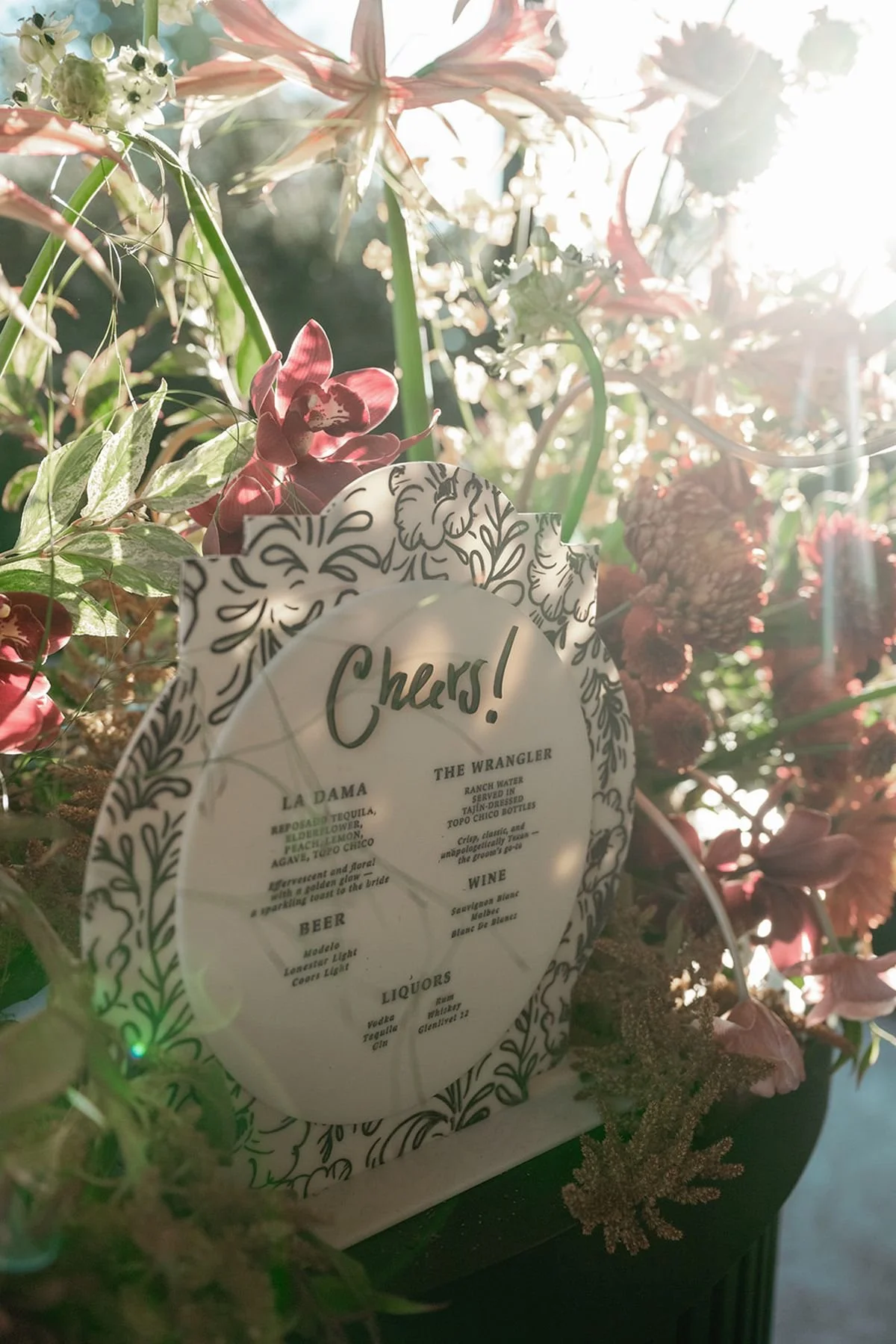 Custom bar menu with “cheers”, specialty cocktails, beer, wine, liquors, surrounded by lush luxury florals in fall colors of plum, burgundy, pink and light streaming in