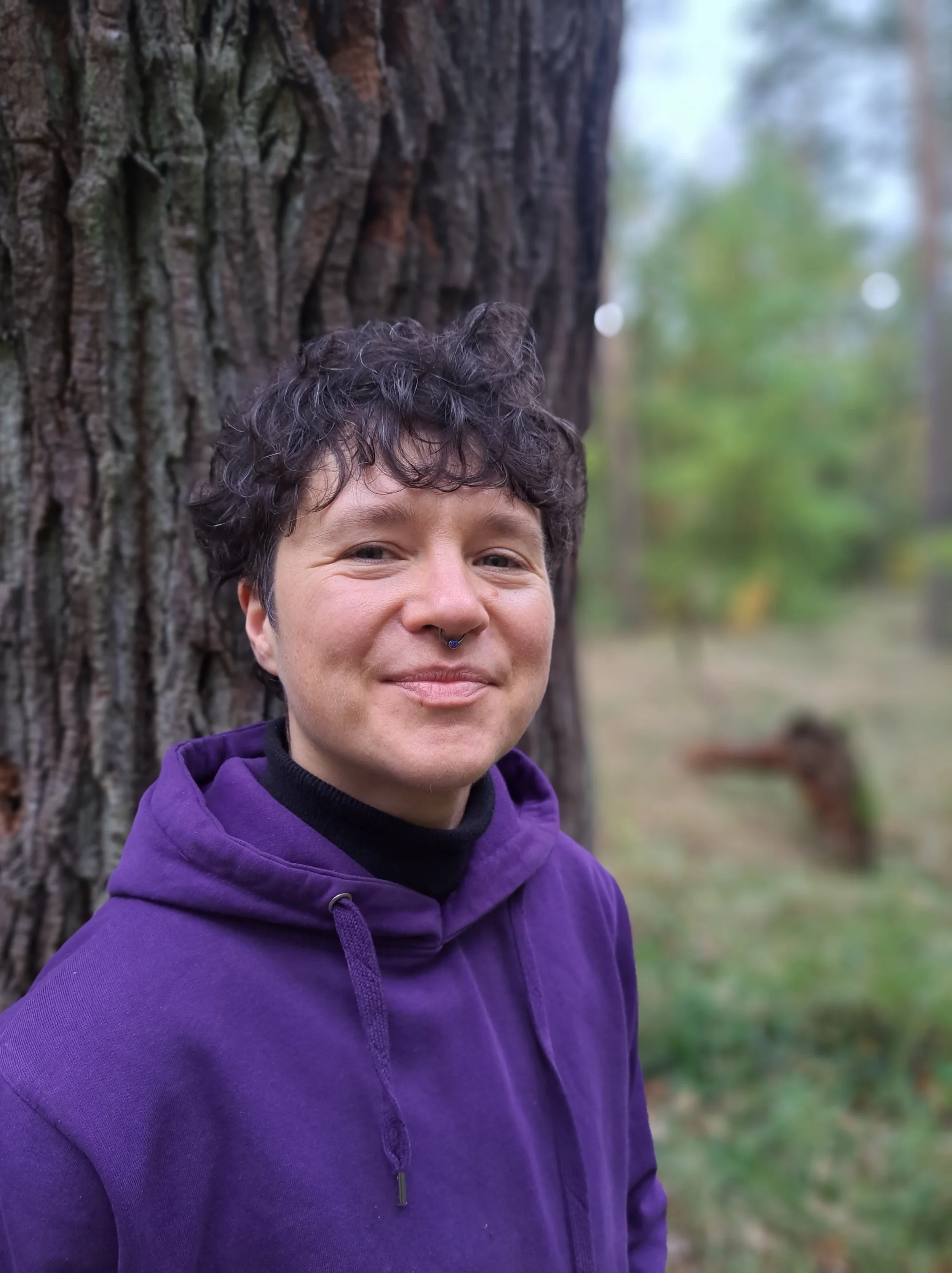 Junge Person mit kurzem, lockigem Haar und Piercing in der Nase, trägt einen lila Hoodie, steht vor einem Baum im Wald.