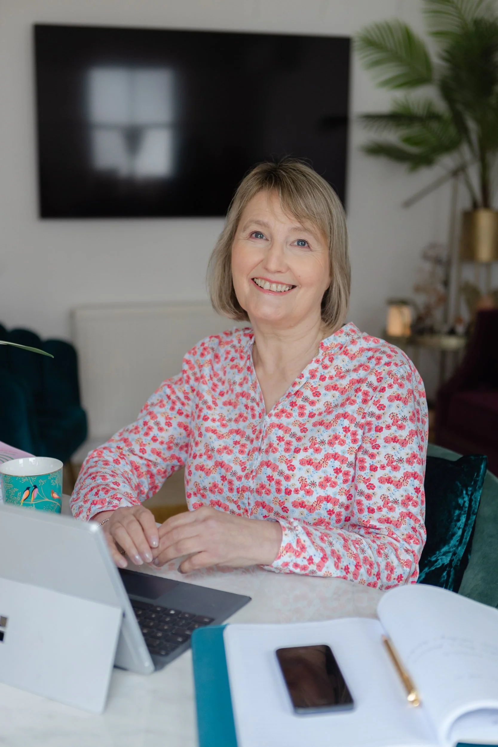 A woman with short blonde hair and a floral blouse smiling while sitting at a desk with a laptop, a mug, a notebook, and a smartphone in a well-lit room with a television and indoor plants in the background.