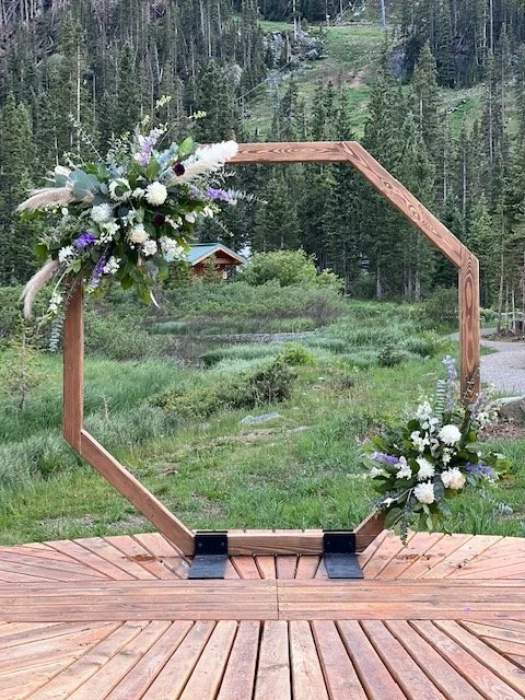 Wooden hexagonal arch decorated with white, purple, and green flowers in an outdoor setting with trees and a grassy area in the background.