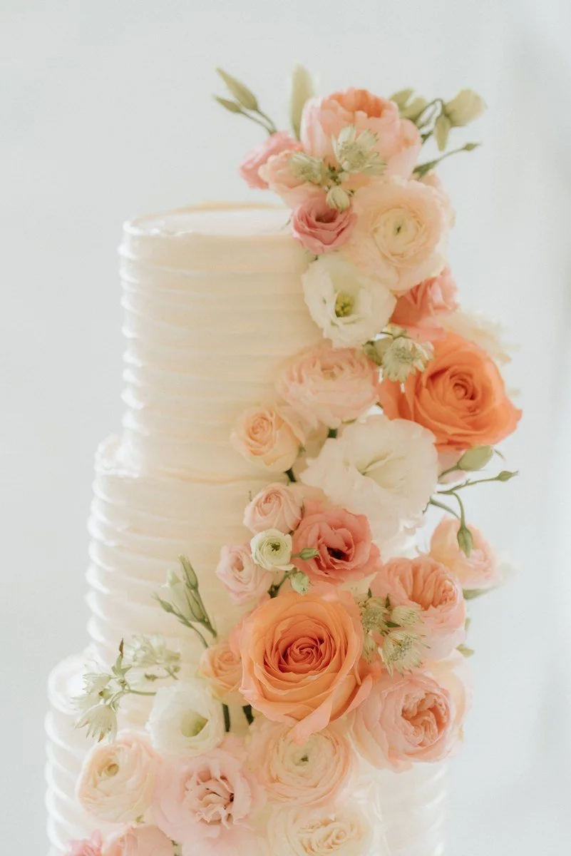 A tall, white, textured wedding cake decorated with a cascading arrangement of peach and white roses and lisianthus flowers.