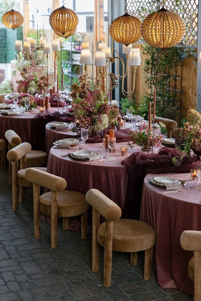 A decorated dining area with tables covered in pinkish-purple tablecloths, floral centerpieces, candle holders, and candles, illuminated by hanging woven pendant lights and wall lamps.