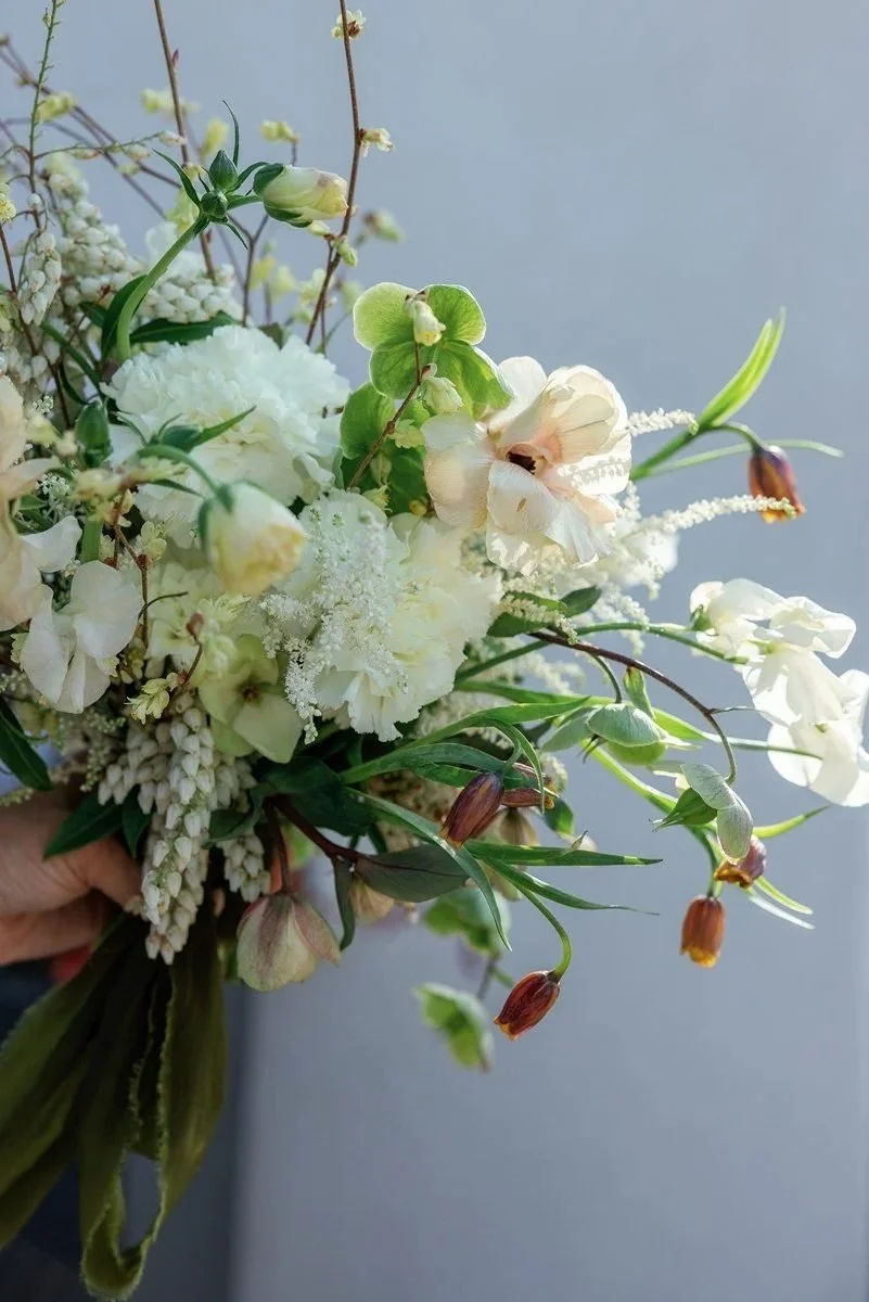 A bouquet of white and cream-colored flowers with green leaves and stems, held in a person's hand against a light gray background.
