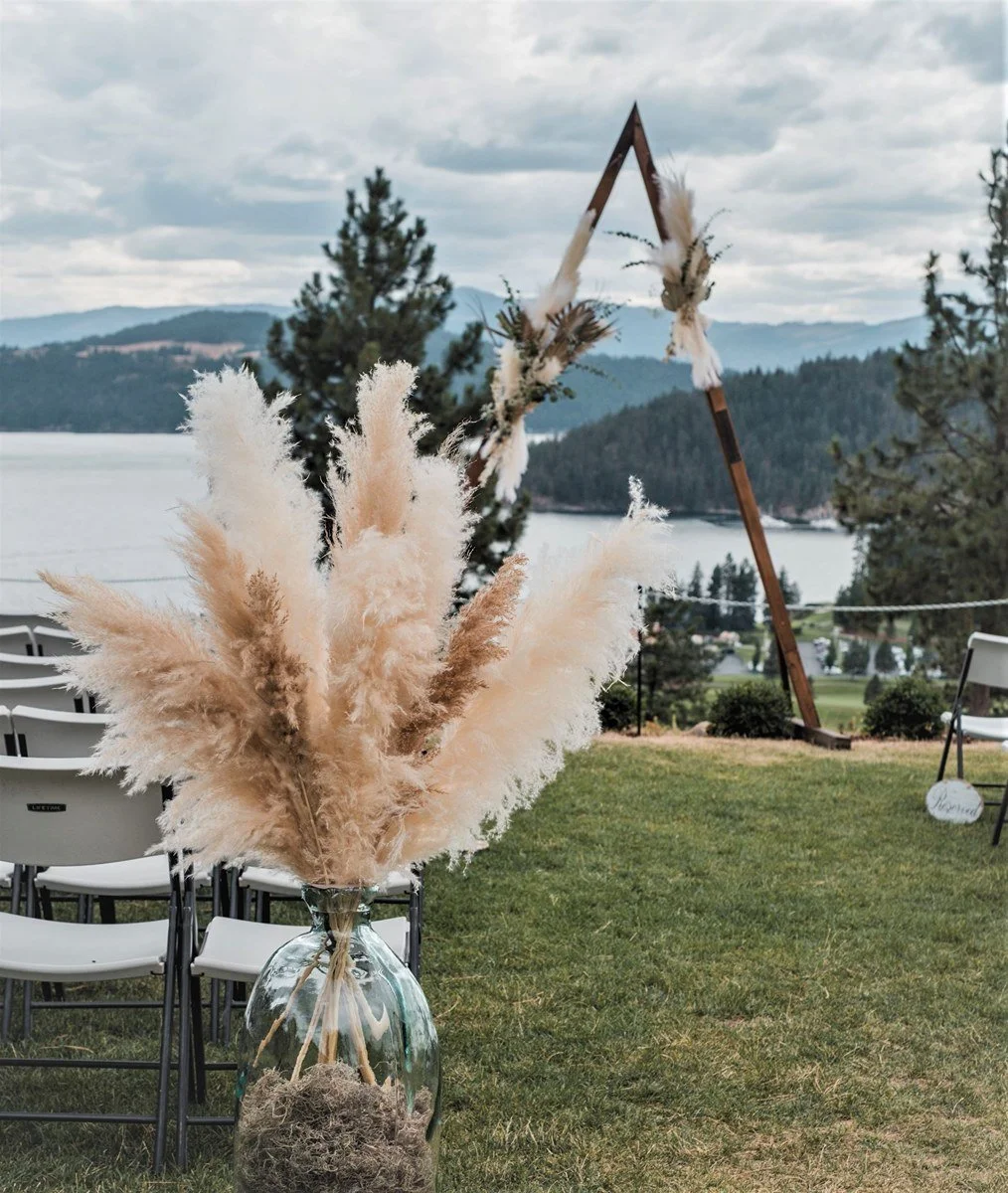 A large glass vase with pampas grass arranged inside, placed on grass with chairs in the background, overlooking a lake and mountains, with a decorative wooden triangle structure nearby.