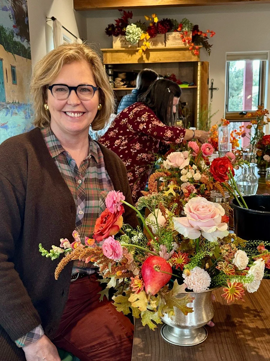 A smiling woman with glasses and earrings sitting at a table with a large flower arrangement in a silver vase, in a cozy room decorated with autumn-themed flowers and decorations.
