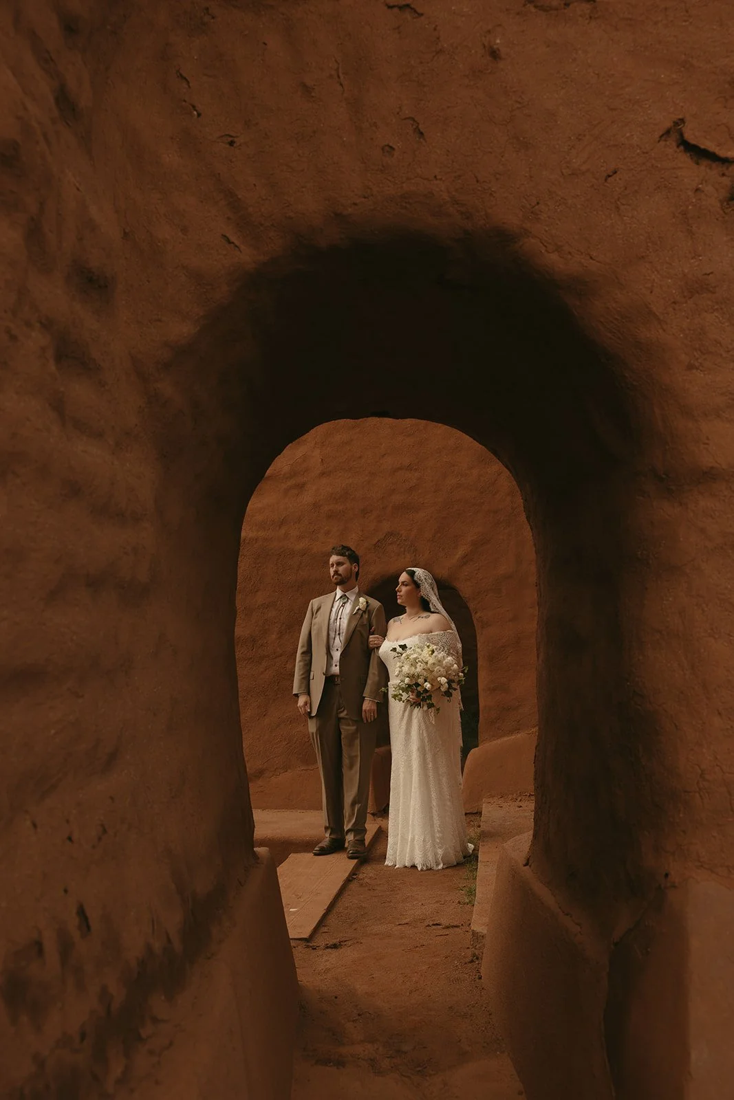A bride and groom standing in a narrow, arched sandstone passageway during their wedding. The bride is holding a bouquet and wearing a lace wedding dress with a veil, while the groom is in a beige suit with a boutonnière.