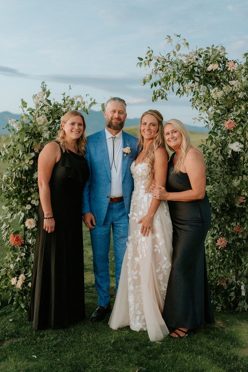 A wedding photo of four people, including a bride in a white lace gown, a groom in a blue suit, and two women in black dresses, standing outdoors with greenery and a floral arch.