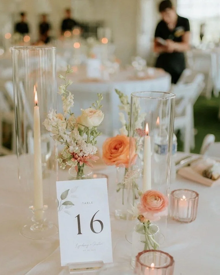 Elegant table centerpiece with white and peach flowers, tall glass candle holders with lit candles, and a table number card reading '16' at a wedding reception.