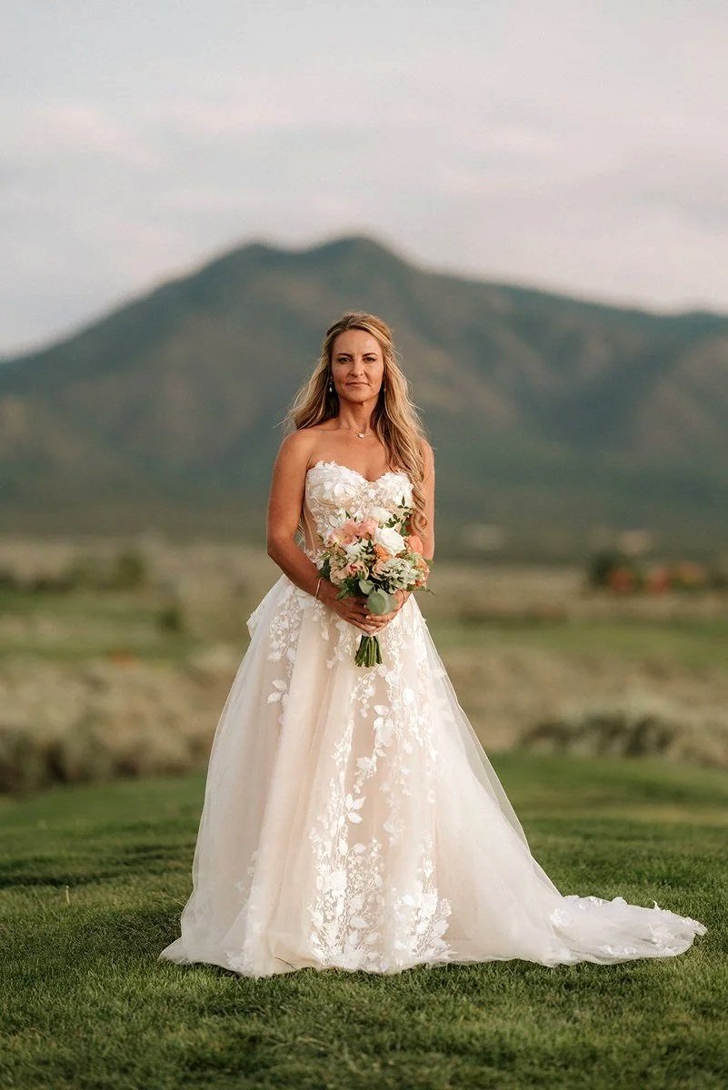 A woman in a strapless wedding dress holding a bouquet of flowers, standing outdoors with mountains in the background.