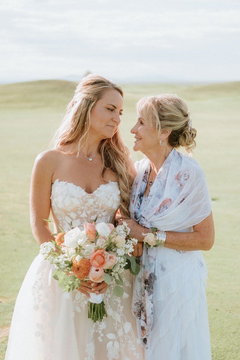 A woman in a white wedding dress holding a bouquet of flowers, looking at an older woman in a white floral shawl, both smiling and standing outdoors on a grassy field.