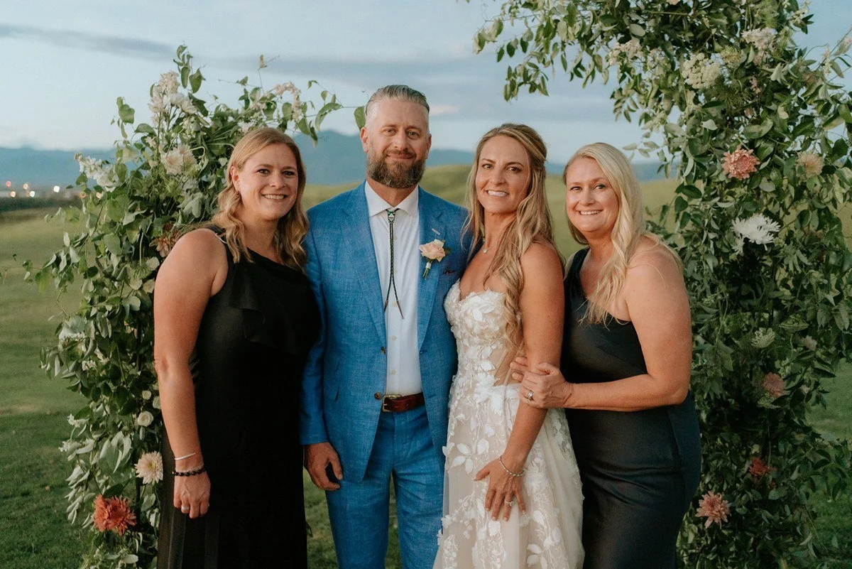 Four people standing outdoors in front of a floral arch at a wedding, with a scenic landscape in the background. The group includes a man in a blue suit and three women in black and white dresses.