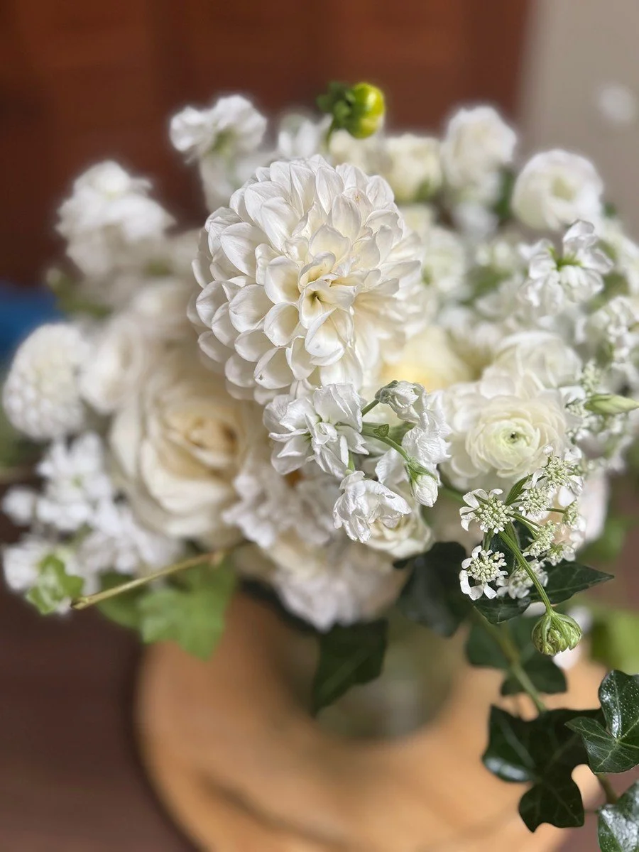 A bouquet of white flowers including dahlias, roses, and lilies, with green leaves in a clear glass vase on a wooden surface.