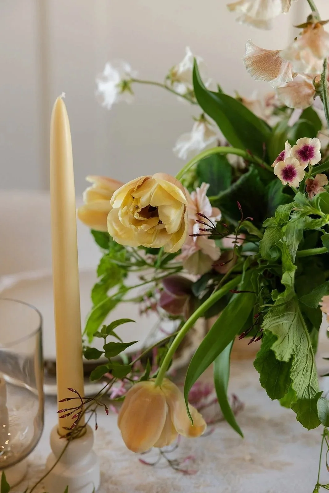 A close-up of a floral arrangement with pastel-colored tulips and other flowers next to a tall, cream-colored taper candle and a glass of water.