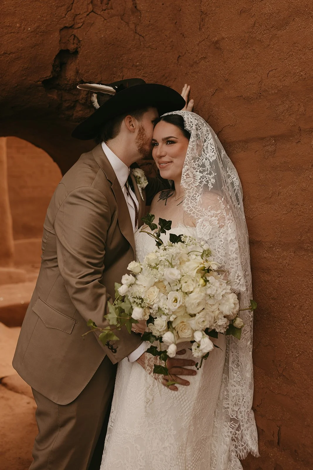 A bride and groom dressed in wedding attire standing close together, with the groom whispering in the bride's ear. The bride is holding a large bouquet of white flowers and wearing a lace veil, while the groom is wearing a brown suit and a black hat 