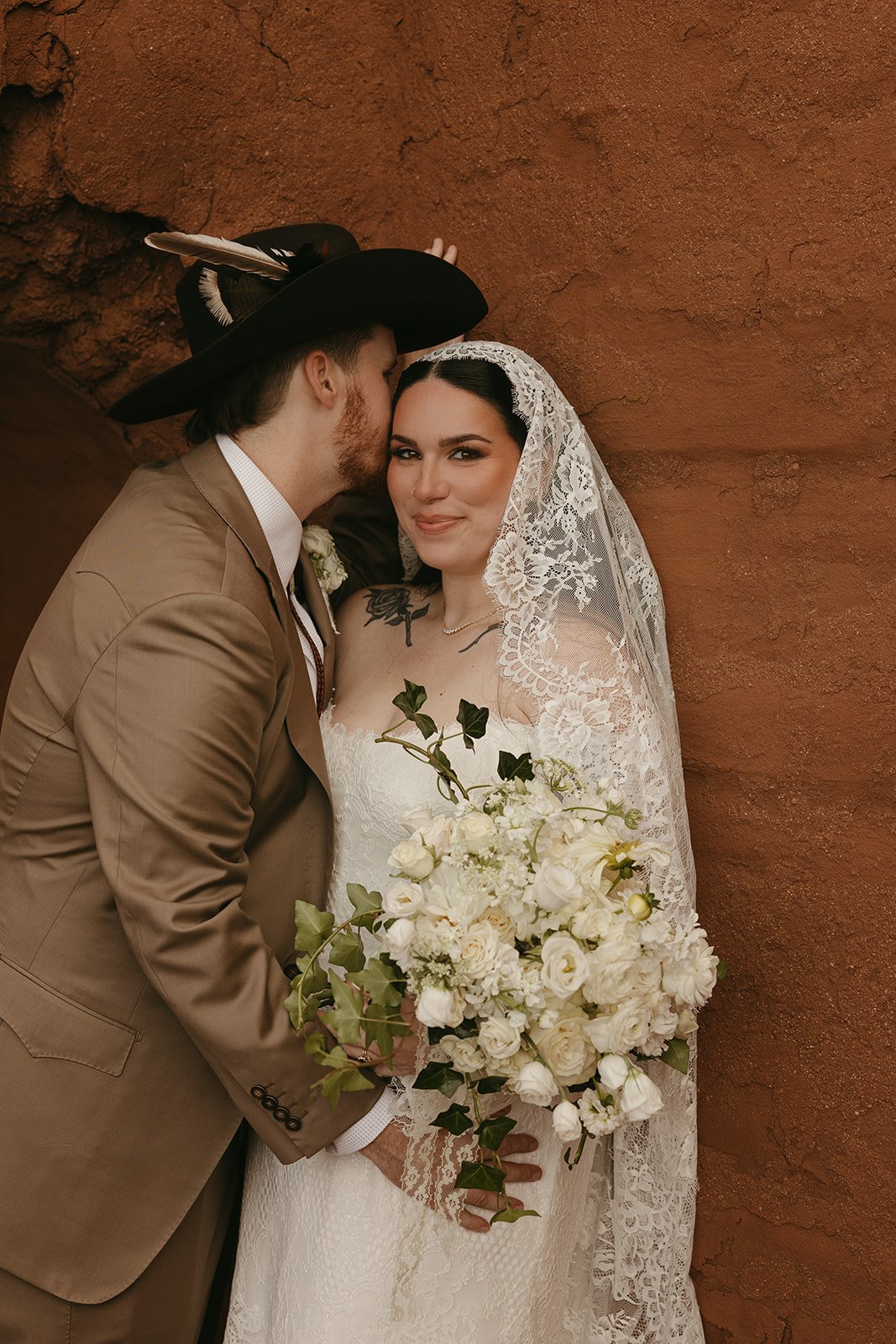 A bride and groom in vintage wedding attire, standing against a textured brick wall. The groom, with reddish hair and beard, is wearing a tan suit and a large black hat with a feather. The bride, with dark hair and a lace veil, is wearing a white lac