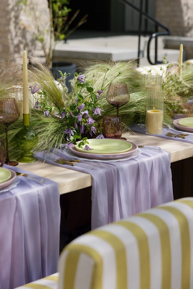 A beautifully decorated outdoor dining table with pastel-colored plates, purple glasses, and a floral centerpiece with purple flowers and greenery, set with candles and gold flatware.