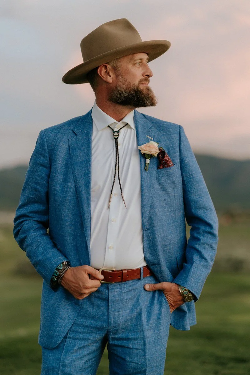 Profile of a man with a beard wearing a light brown wide-brimmed hat, a blue suit, white shirt, patterned bolo tie, and a pink rose boutonnière, standing outdoors during sunset.