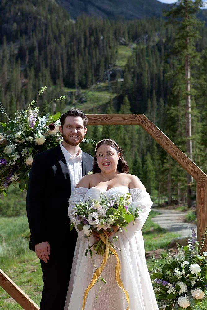A newlywed couple standing outdoors in front of a wooden arch decorated with flowers, with a forested mountain landscape in the background.