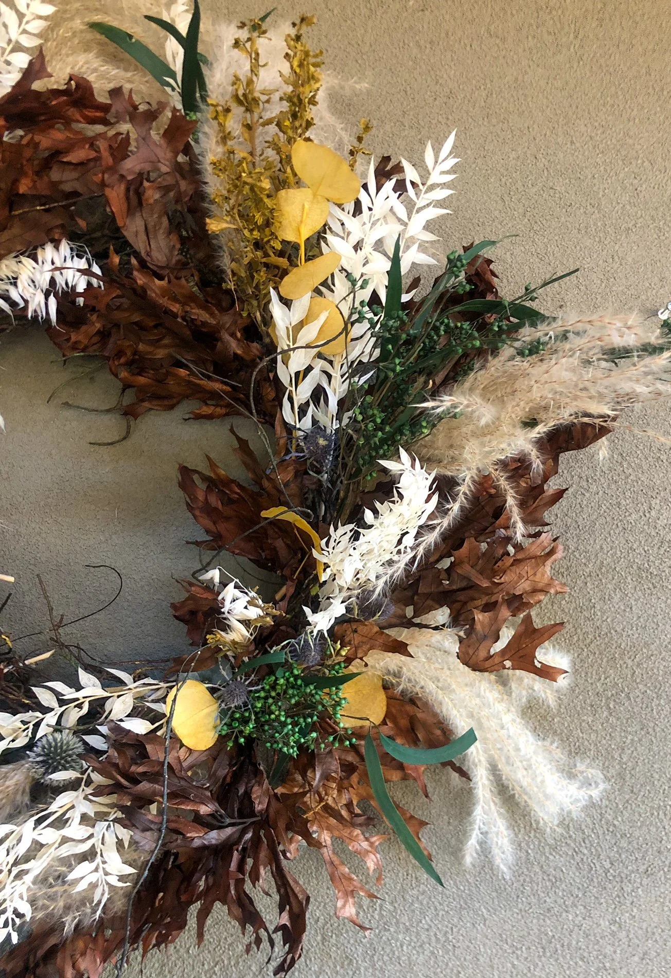 A fall-themed decorative arrangement featuring brown dried leaves, white and yellow artificial flowers, green berries, and fluffy pampas grass on a neutral background.