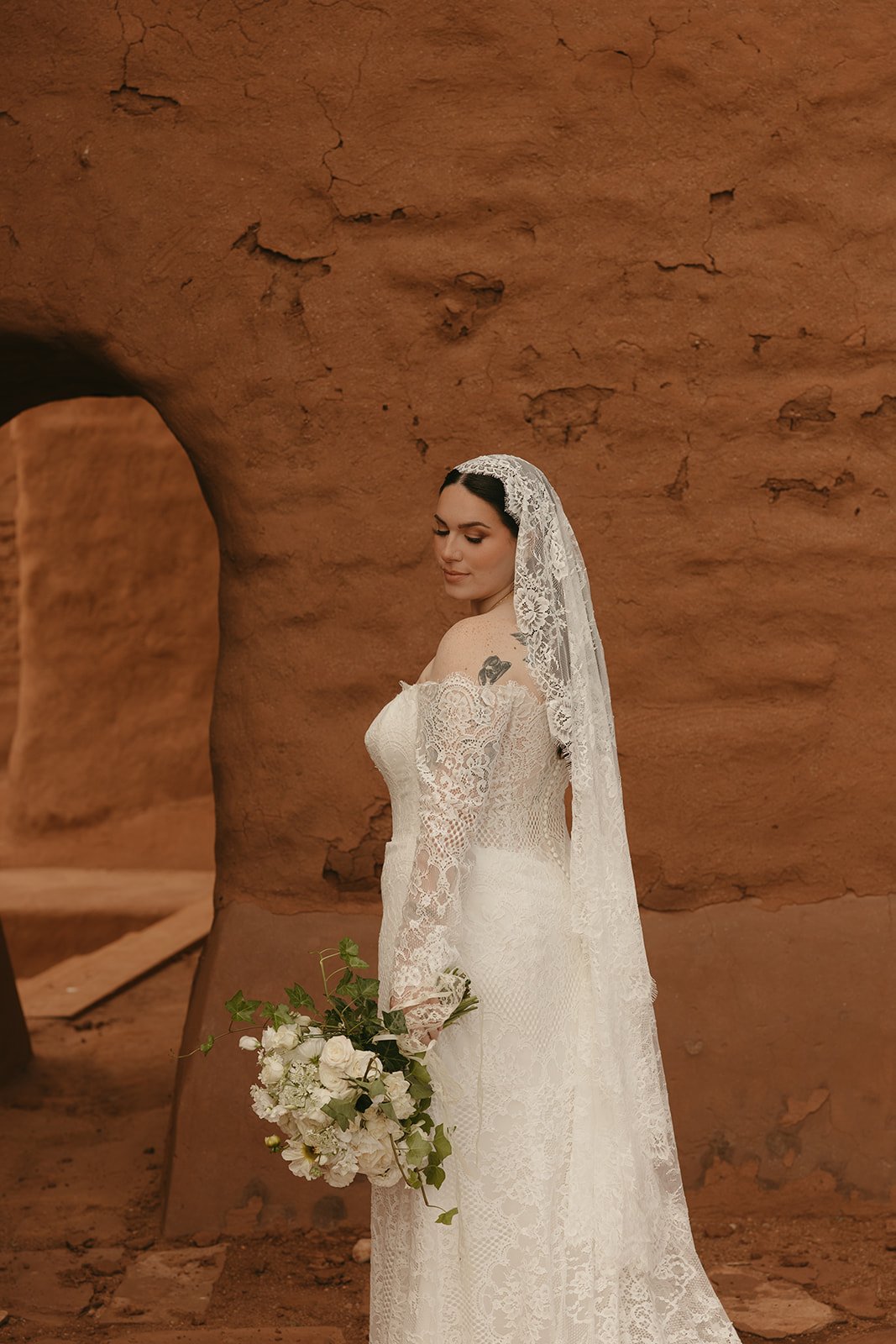 A woman in a lace wedding dress with a lace veil, holding a bouquet of white flowers, standing against a desert sand-colored rock formation.