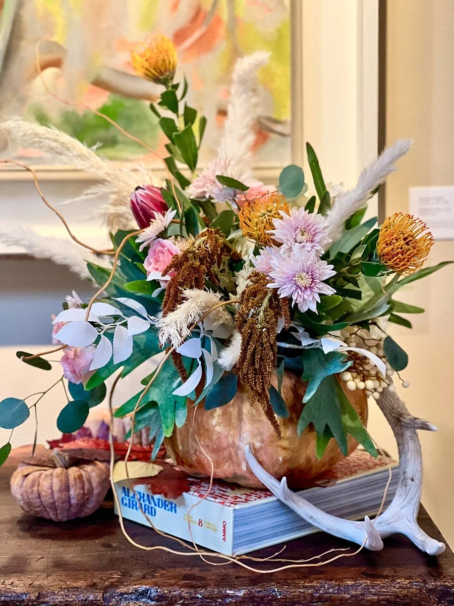 A floral arrangement with various colorful flowers, green leaves, and decorative elements in a round vessel, placed on a book and a small pumpkin on a wooden surface.