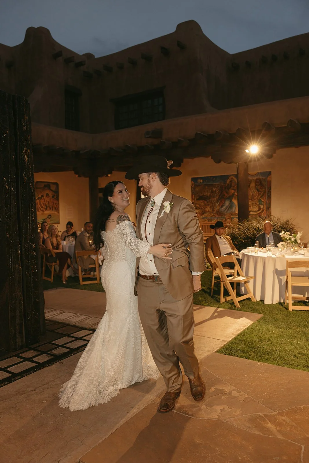 A bride and groom sharing a dance at their wedding reception in a rustic outdoor setting, with guests seated at tables in the background.