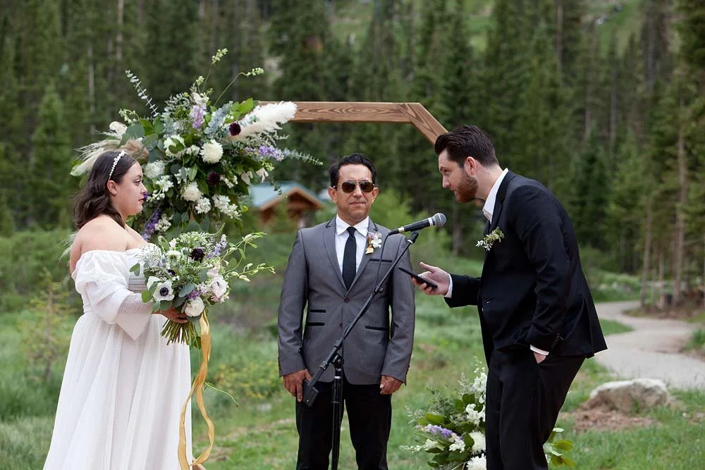 A wedding ceremony outdoors with a bride holding a bouquet, a groom reading vows, and an officiant standing between them in a forest setting.