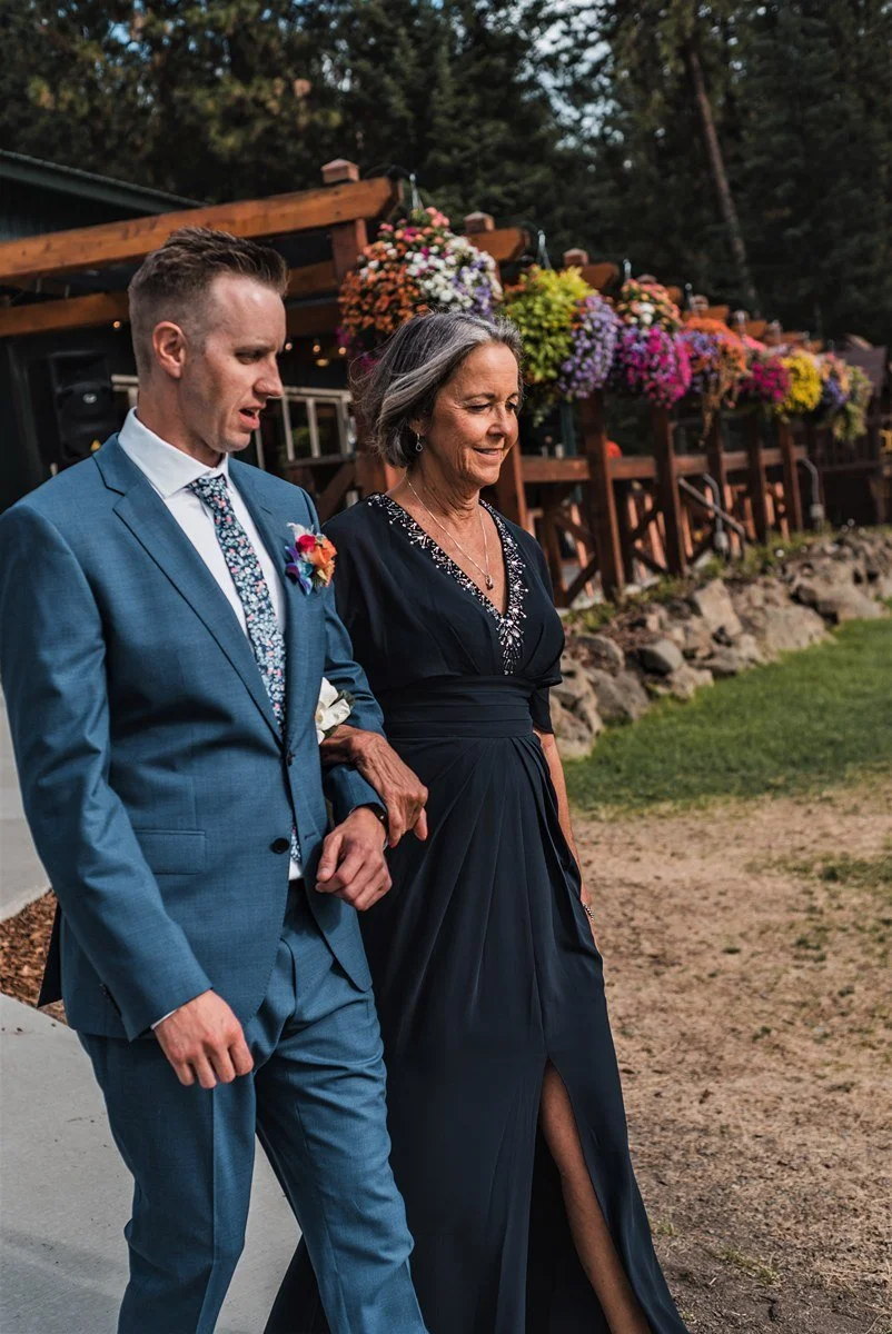 A young man in a blue suit and floral tie walks arm-in-arm with an older woman in a black evening gown outdoors during a celebration, with colorful flowers in the background.