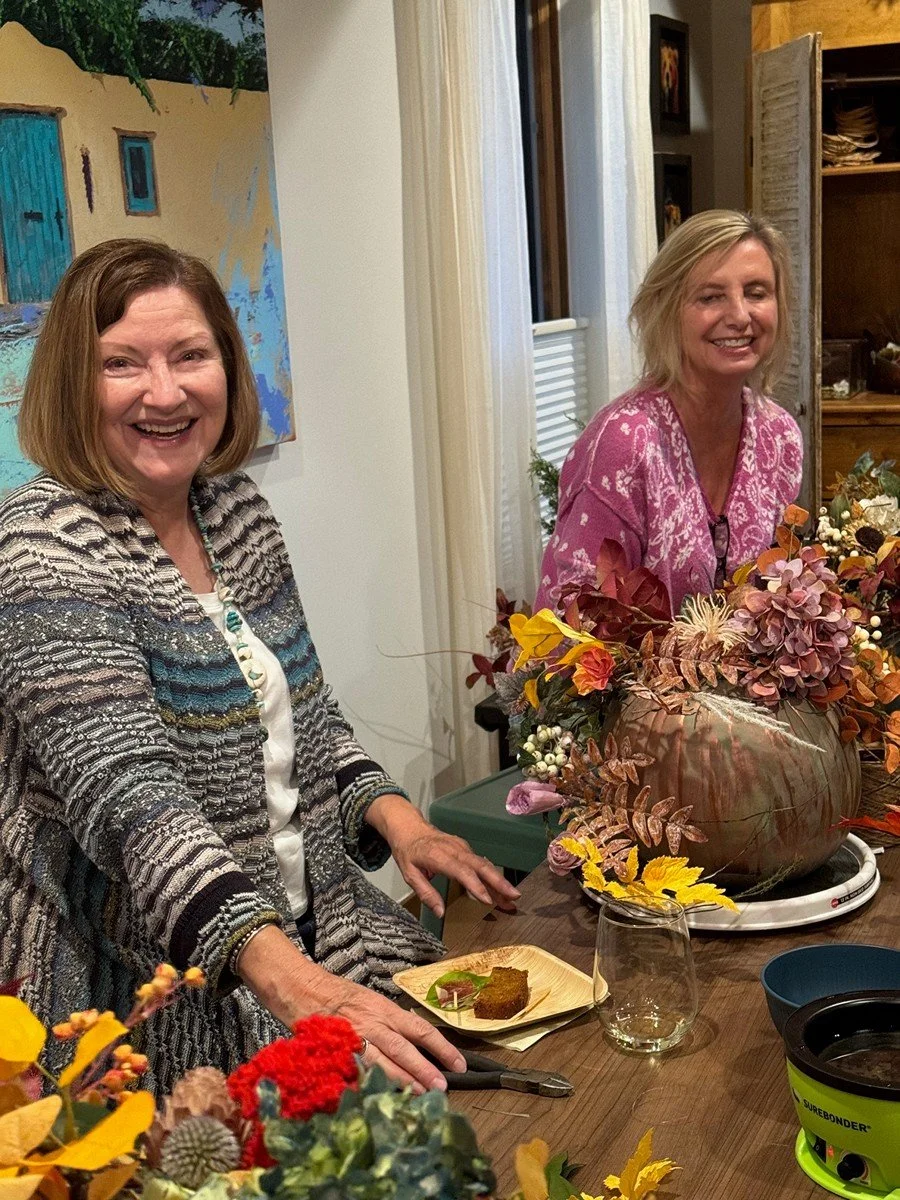Two women smiling at a dining table decorated with fall floral arrangement in a large vase, with food and glasses on the table, inside a cozy room.