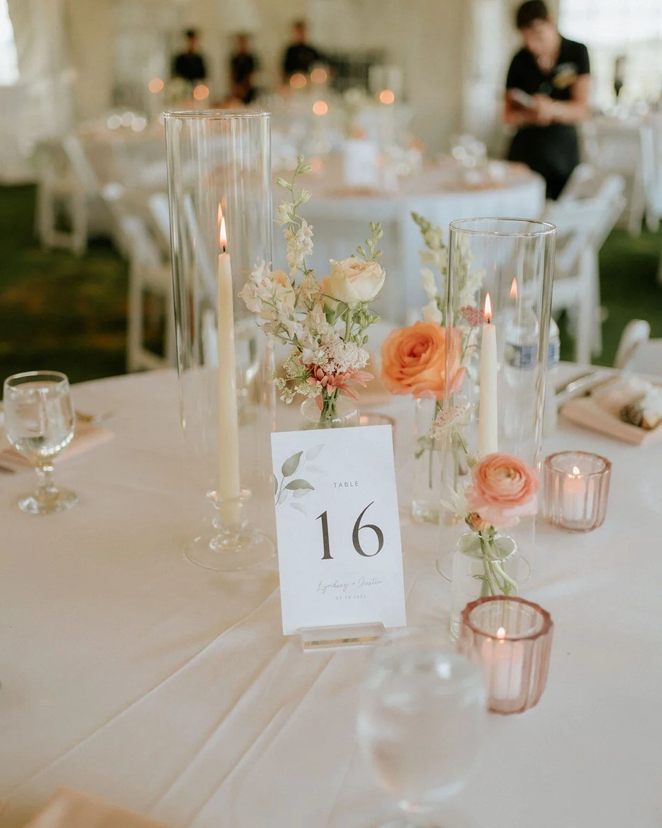 Elegant table centerpiece with white and peach flowers, tall glass candle holders with lit candles, and a table number card reading '16' at a wedding reception.