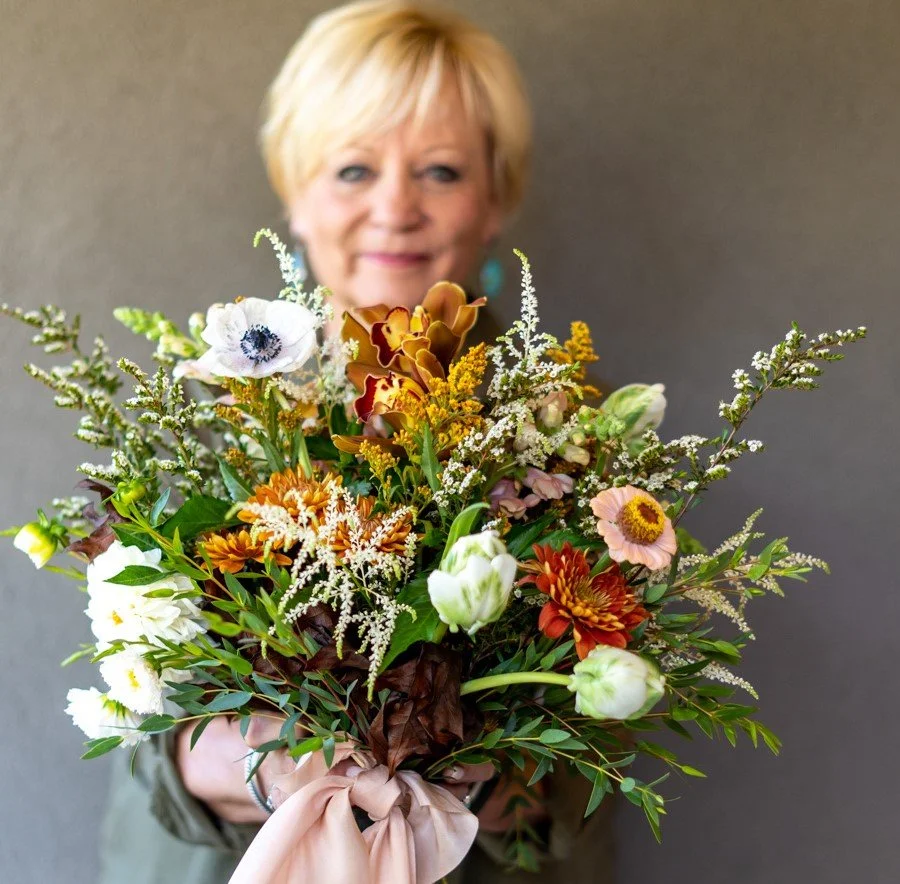 A woman with short blonde hair holding a large bouquet of colorful flowers, with a neutral gray background.